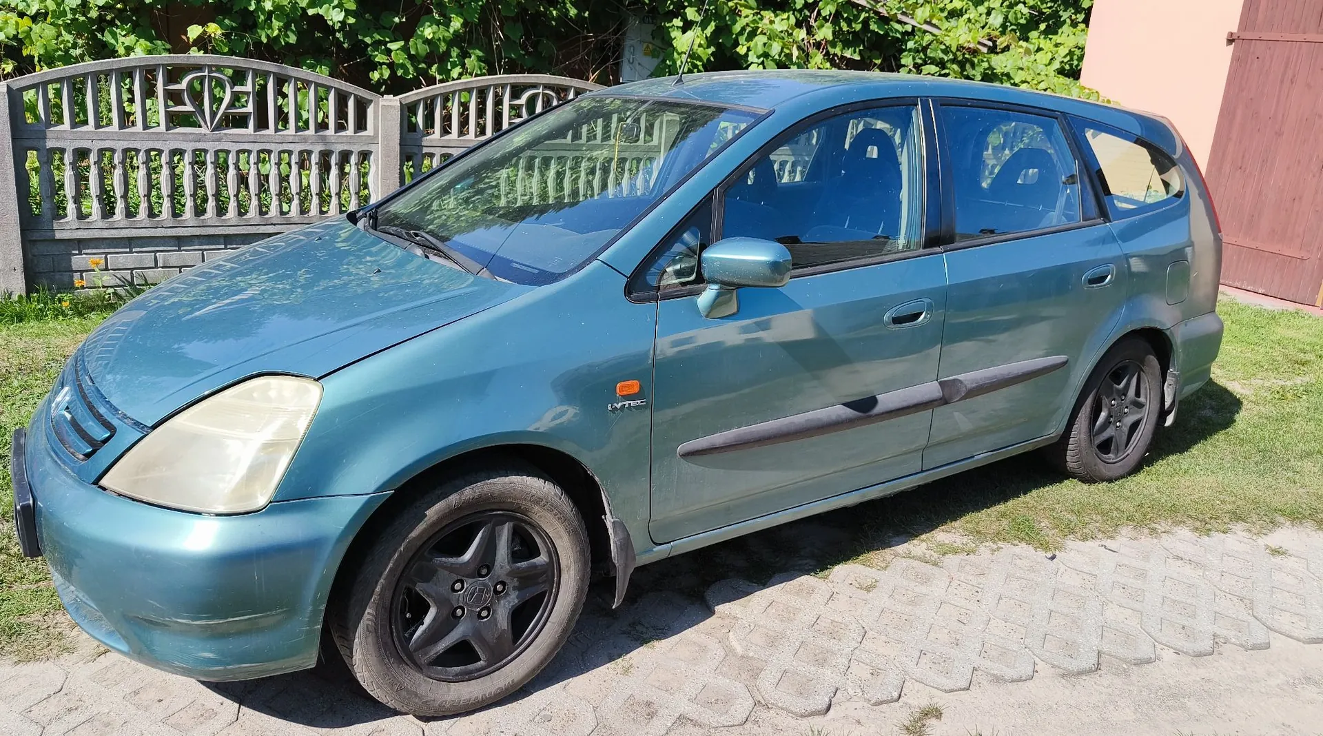 Niebieski samochód typu kombi zaparkowany na podjeździe z kostki brukowej. Pojazd jest widoczny z profilu, z przodu widać zmatowiałe reflektory. Na przednim błotniku znajduje się mały napis 'VTEC'. W tle widoczny jest ozdobny betonowy płot oraz zielone krzewy i fragment ściany budynku w kolorze beżowym.