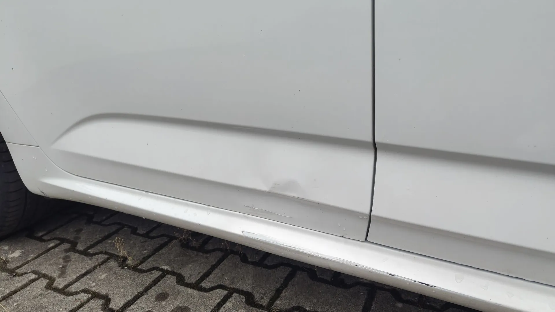 Zbliżenie na dolną część białego nadwozia samochodu. Na boku auta, wzdłuż dolnej krawędzi drzwi, widoczne jest wgniecenie oraz kilka drobnych zarysowań lakieru. Samochód zaparkowany jest na szarej kostce brukowej.