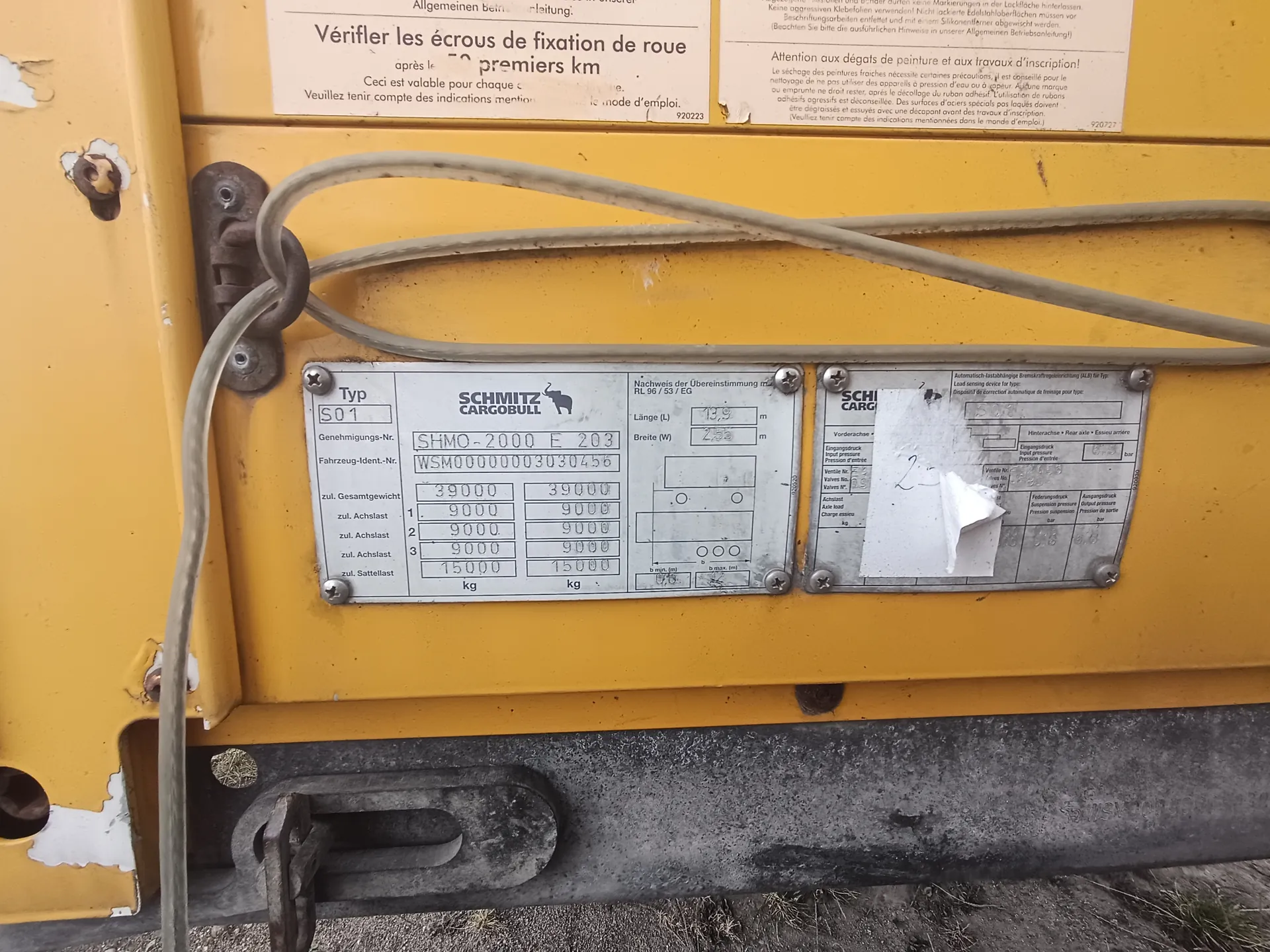 Zbliżenie na żółtą powierzchnię pojazdu z metalowymi tabliczkami identyfikacyjnymi. Lewa tabliczka zawiera napisy: 'Typ S01', 'SCHMITZ CARGOBULL' z logo słonia, 'Genehmigungs-Nr. SHMO-2000 E 203', 'Fahrzeug-Ident.-Nr. WSM00000003030456', 'zul. Gesamtgewicht 39000 kg', 'zul. Achslast 1. 9000 kg', '2. 9000 kg', '3. 15000 kg'. Prawa tabliczka częściowo zakryta białym naklejką, widoczny fragment 'SCHI CARG'. Nad tabliczkami dwa dokumenty z tekstem w języku francuskim i niemieckim, m.in. 'Vérifier les écrous de fixation de roue après les 50 premiers km' oraz 'Attention aux dégâts de peinture et aux travaux d'inscription!'. Na żółtej powierzchni widoczne są szare przewody, zardzewiałe elementy metalowe i ślady zużycia malowania. W dolnej części metalowy hak i brudna powierzchnia.