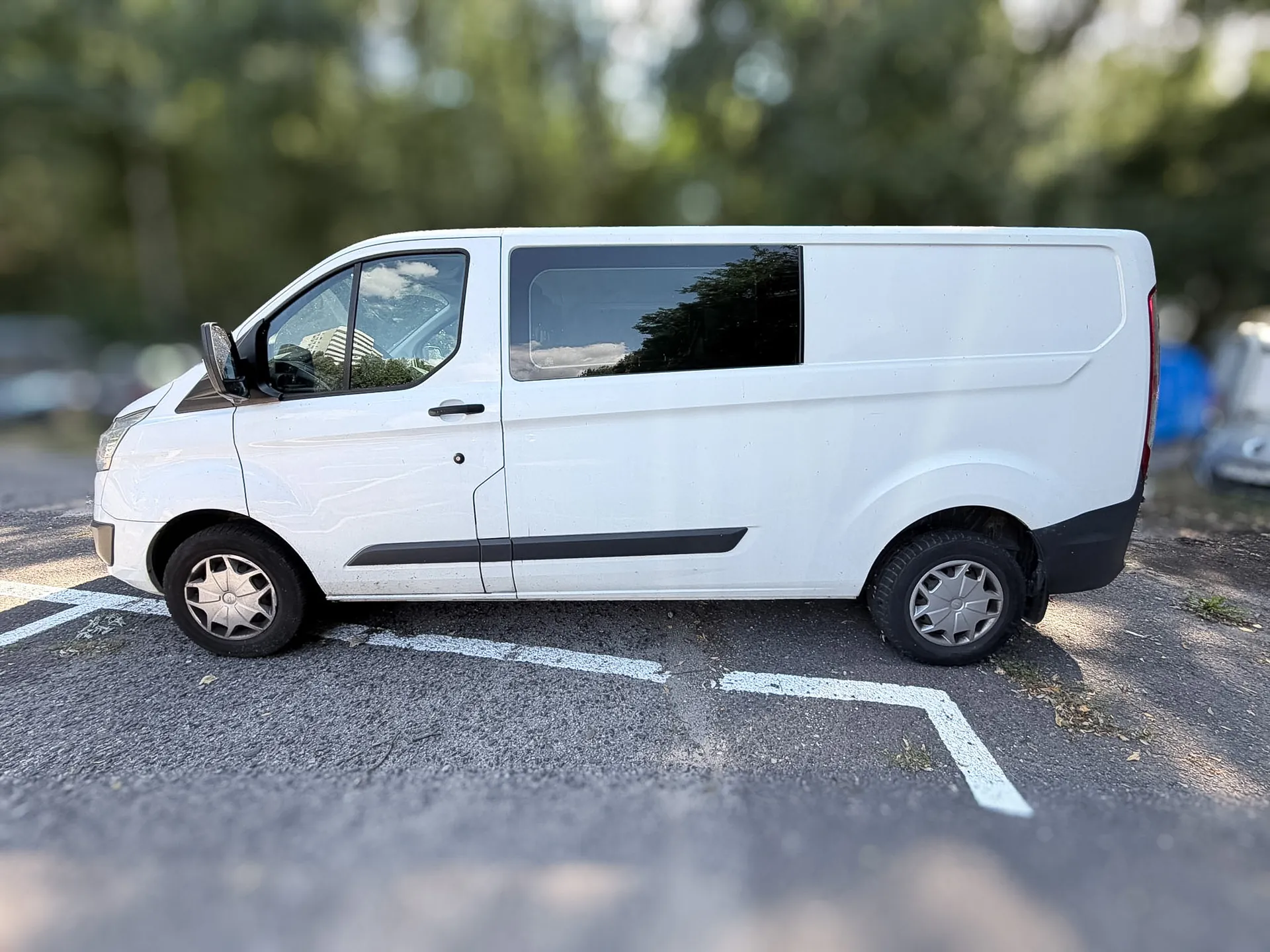 Biały furgon zaparkowany bokiem na asfaltowym parkingu z białymi liniami wyznaczającymi miejsce postojowe. Pojazd ma przyciemniane tylne szyby i czarne detale, takie jak klamki i listwy boczne. W tle widoczne są rozmyte zielone drzewa.