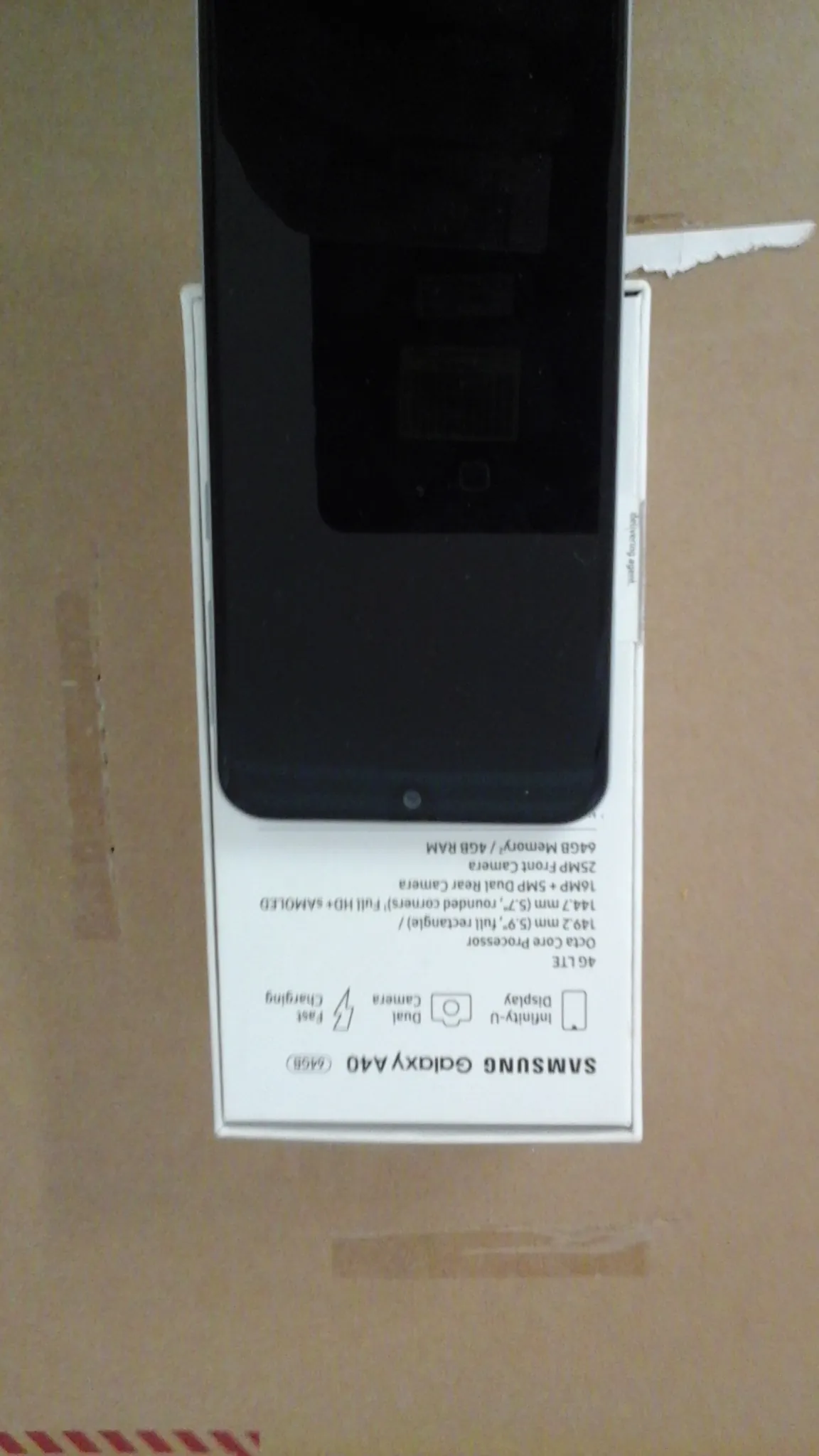 Białe pudełko Samsung Galaxy A40 z napisem 64GB, przedstawiające czarny smartfon. Na pudełku widoczne informacje: Infinity-U Display, Dual Camera, Fast Charging, 4G LTE, Octa Core Processor, 149,2 mm (5,9", full rectangle)/144,7 mm (5,7", rounded corners) Full HD+ sAMOLED, 16MP + 5MP Dual Rear Camera, 25MP Front Camera, 64GB Memory / 4GB RAM. Tło stanowi brązowa powierzchnia kartonowa z czerwonymi pionowymi paskami po lewej stronie oraz fragmentami taśmy.