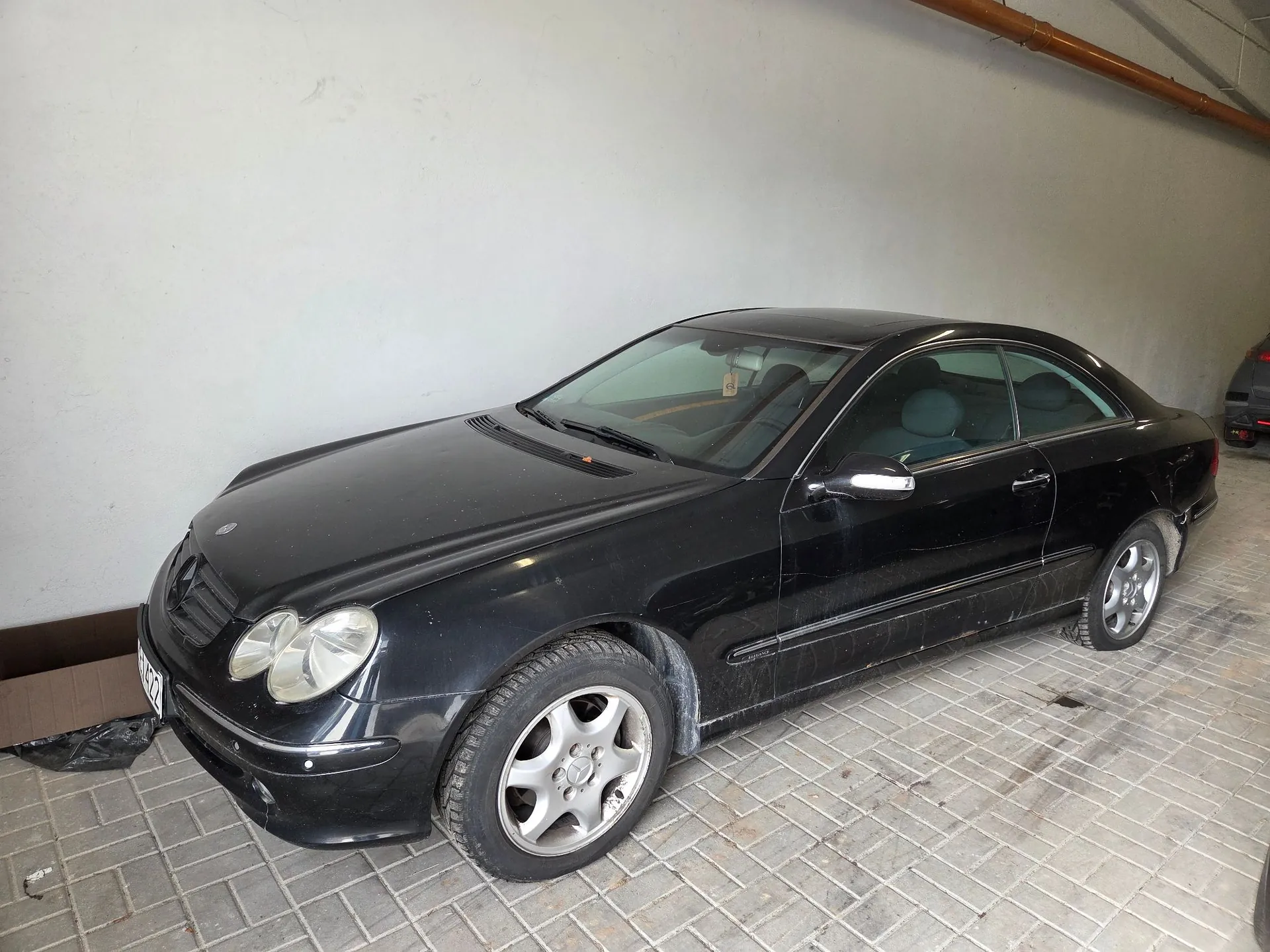 Czarny samochód osobowy marki Mercedes-Benz w nadwoziu coupe, zaparkowany w garażu na posadzce z szarej kostki brukowej. Pojazd jest zakurzony i ustawiony pod kątem, ukazując przód oraz prawy bok auta.