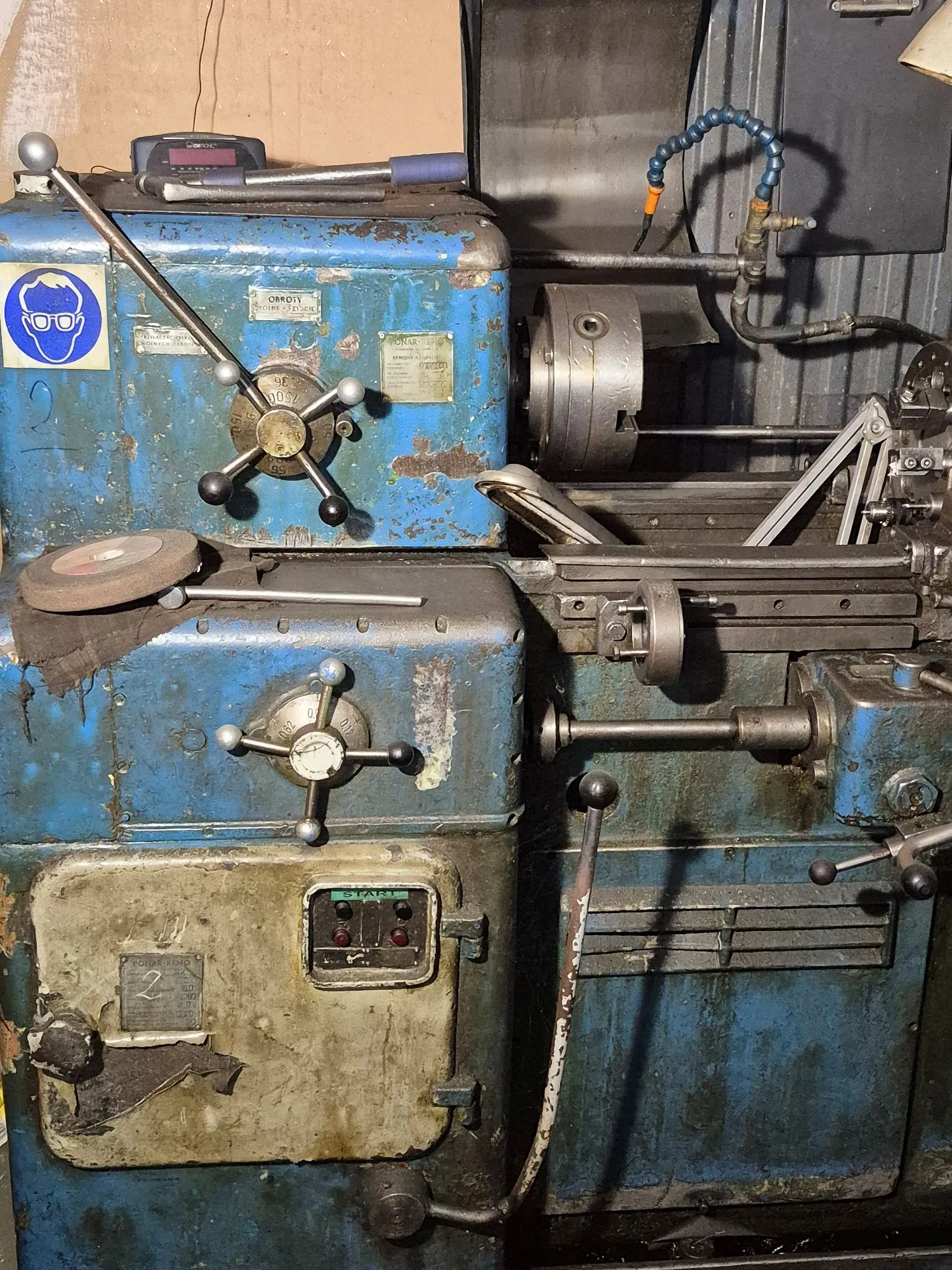 Stara, zużyta maszyna przemysłowa w kolorze niebieskim z widocznymi śladami rdzy i odpryskami farby. Urządzenie posiada liczne metalowe pokrętła, dźwignie oraz tabliczki znamionowe. Na górze znajduje się cyfrowy miernik oraz niebieski przedmiot przypominający narzędzie. W dolnej części widoczny jest panel sterowania z zielonym przyciskiem START i dwiema czerwonymi lampkami kontrolnymi.