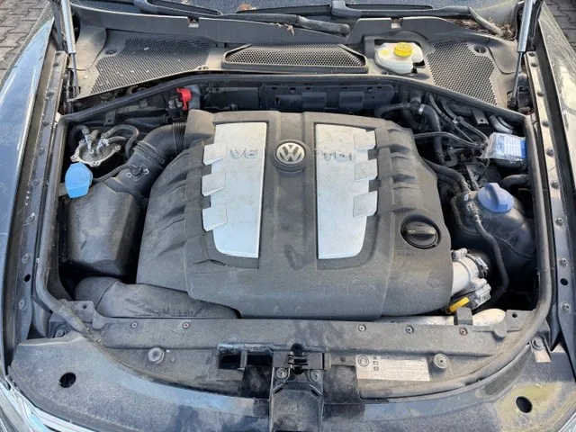 Otwarty maska samochodu marki Volkswagen z widocznym silnikiem V6. Na pokrywie silnika umieszczono logo VW oraz napisy V6 po obu stronach. Wokół silnika znajdują się różne elementy: niebieska nakrętka po lewej stronie, niebieska nakrętka po prawej stronie, żółta nakrętka w górnej części oraz czarne przewody i zbiorniki. W dolnej części obrazu widoczna jest tabliczka z niewyraźnymi danymi technicznymi. Tło stanowi brukowana powierzchnia.