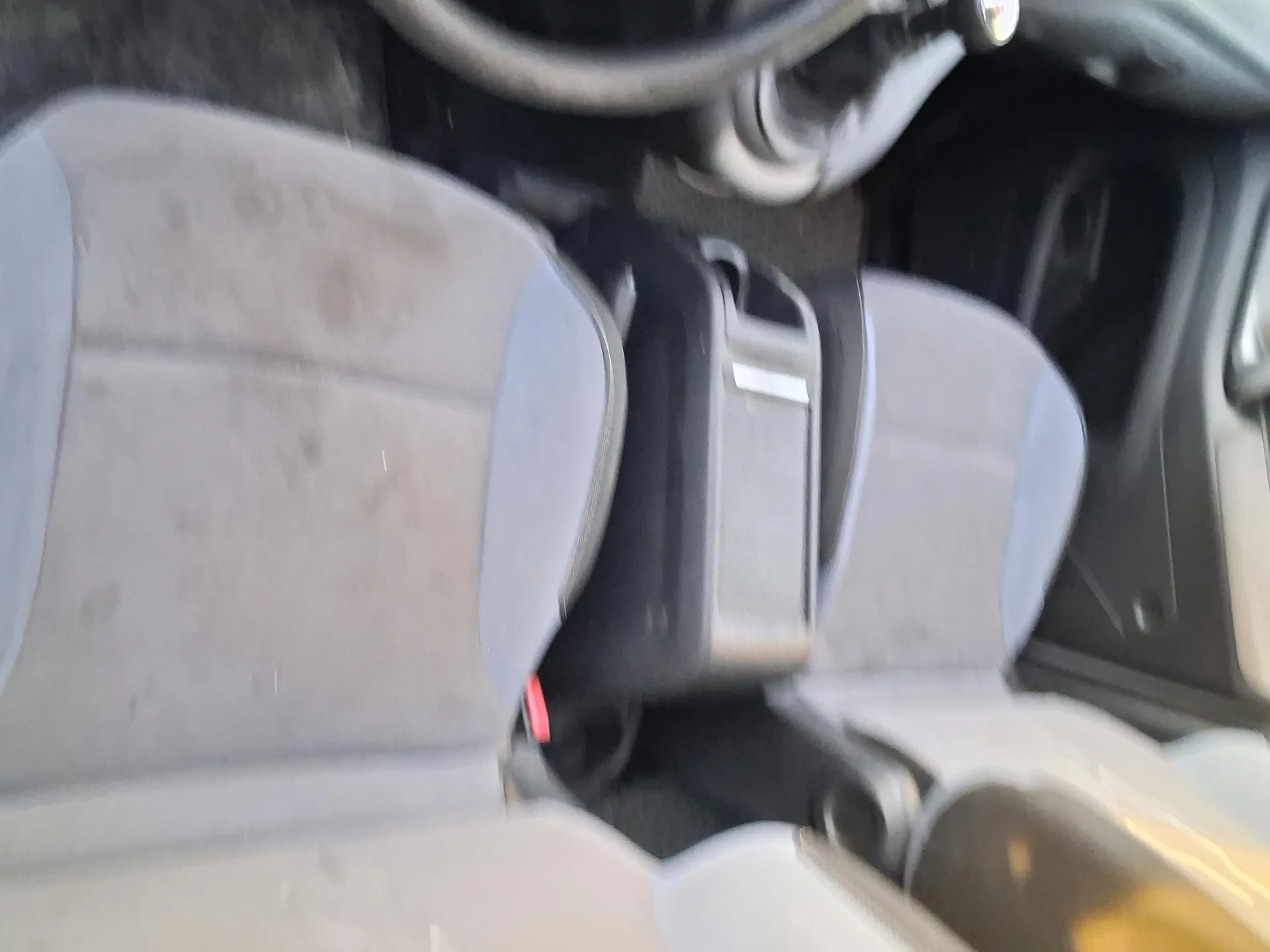 Wnętrze samochodu z widocznymi przódowymi siedzeniami w kolorze szarym, częściowo zabrudzonymi. Między siedzeniami znajduje się centralna konsoleta z dźwignią zmiany biegów po lewej stronie, a także otworem na napoje i przestrzenią na przechowywanie. Widoczne są fragmenty drzwi z uchwytem i czarną podłogą. Na prawo od konsolety znajduje się czerwony zamek pasów bezpieczeństwa.