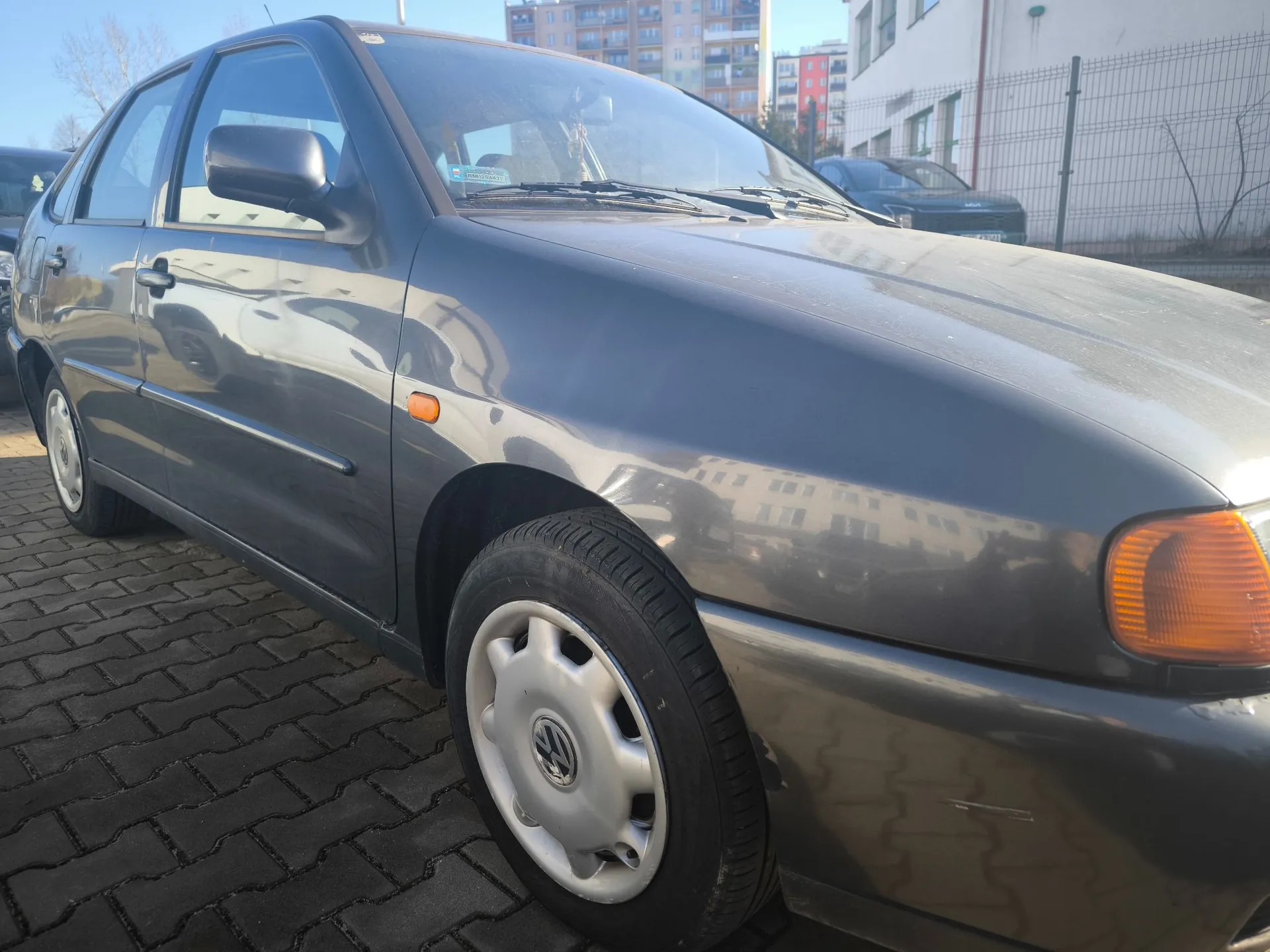 Szary samochód osobowy marki Volkswagen zaparkowany na kostce brukowej. Zdjęcie przedstawia przód i bok pojazdu z perspektywy trzech czwartych. W tle widoczne są inne samochody, ogrodzenie z siatki oraz wielopiętrowe budynki mieszkalne pod jasnym niebem.