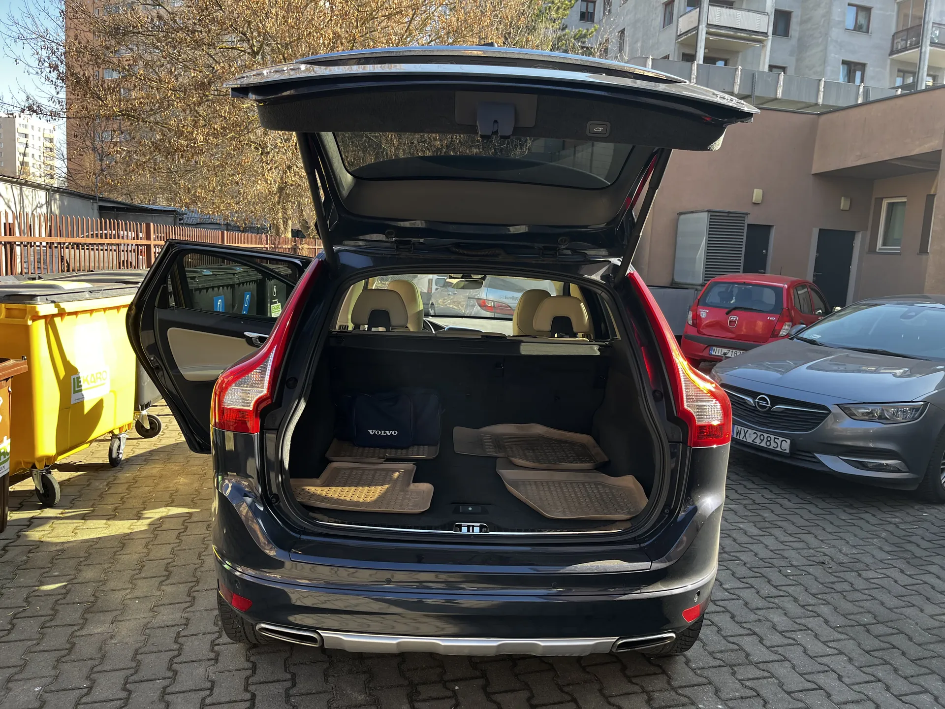 Czarny samochód Volvo z otwartym bagażnikiem i otwartą lewą tylną drzwiami. W bagażniku widoczna niebieska torba z napisem VOLVO oraz brązowe dywaniki. Po lewej stronie żółty pojemnik na śmieci z napisem EKO. W tle szary samochód Opel z tablicą rejestracyjną WX 2985C, czerwony samochód, budynki mieszkalne, suche drzewa i brukowana powierzchnia. Słoneczna pogoda.