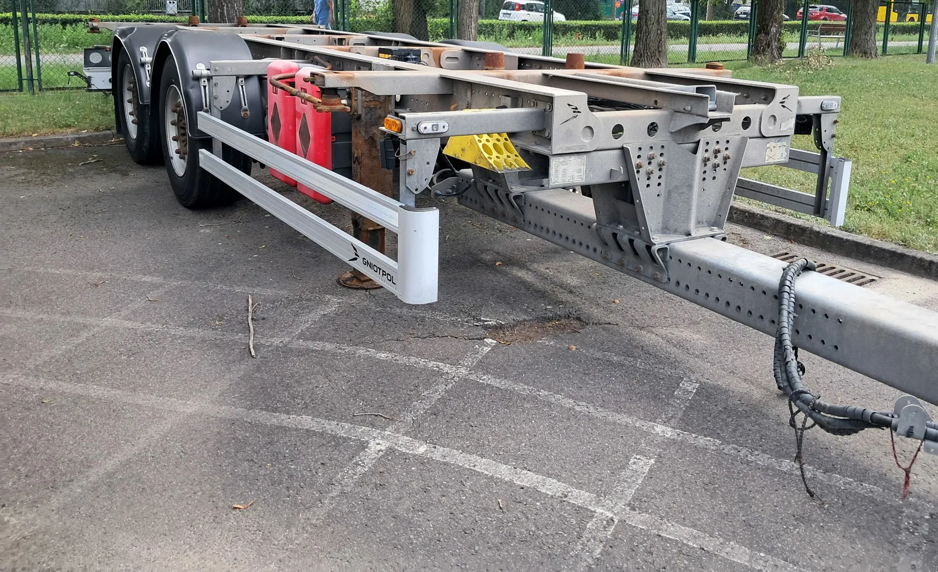 Srebrna, dwuosiowa przyczepa transportowa zaparkowana na asfaltowym placu. Przyczepa posiada metalową ramę, dwa koła z lewej strony oraz czerwone elementy odblaskowe. Na białym panelu boczny widnieje napis „GNIOTPOL”. W tle widać zielone drzewa, ogrodzenie oraz zaparkowane samochody.