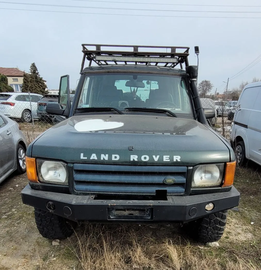 Widok z przodu ciemnozielonego samochodu terenowego marki Land Rover. Pojazd posiada czarny zderzak, dodatkowe oświetlenie LED na dachu zamontowane na metalowym bagażniku oraz terenowe opony. Na masce widnieje biały napis 'LAND ROVER'. W tle widoczne są inne zaparkowane samochody, ogrodzenie z siatki oraz budynki mieszkalne pod zachmurzonym niebem.