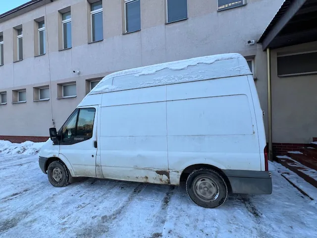 Biały furgon marki Ford Transit zaparkowany na zaśnieżonym podjeździe przed jasnym, wielokondygnacyjnym budynkiem z rzędem okien. Na dachu samochodu znajduje się warstwa śniegu, a dolna część nadwozia jest zabrudzona błotem.