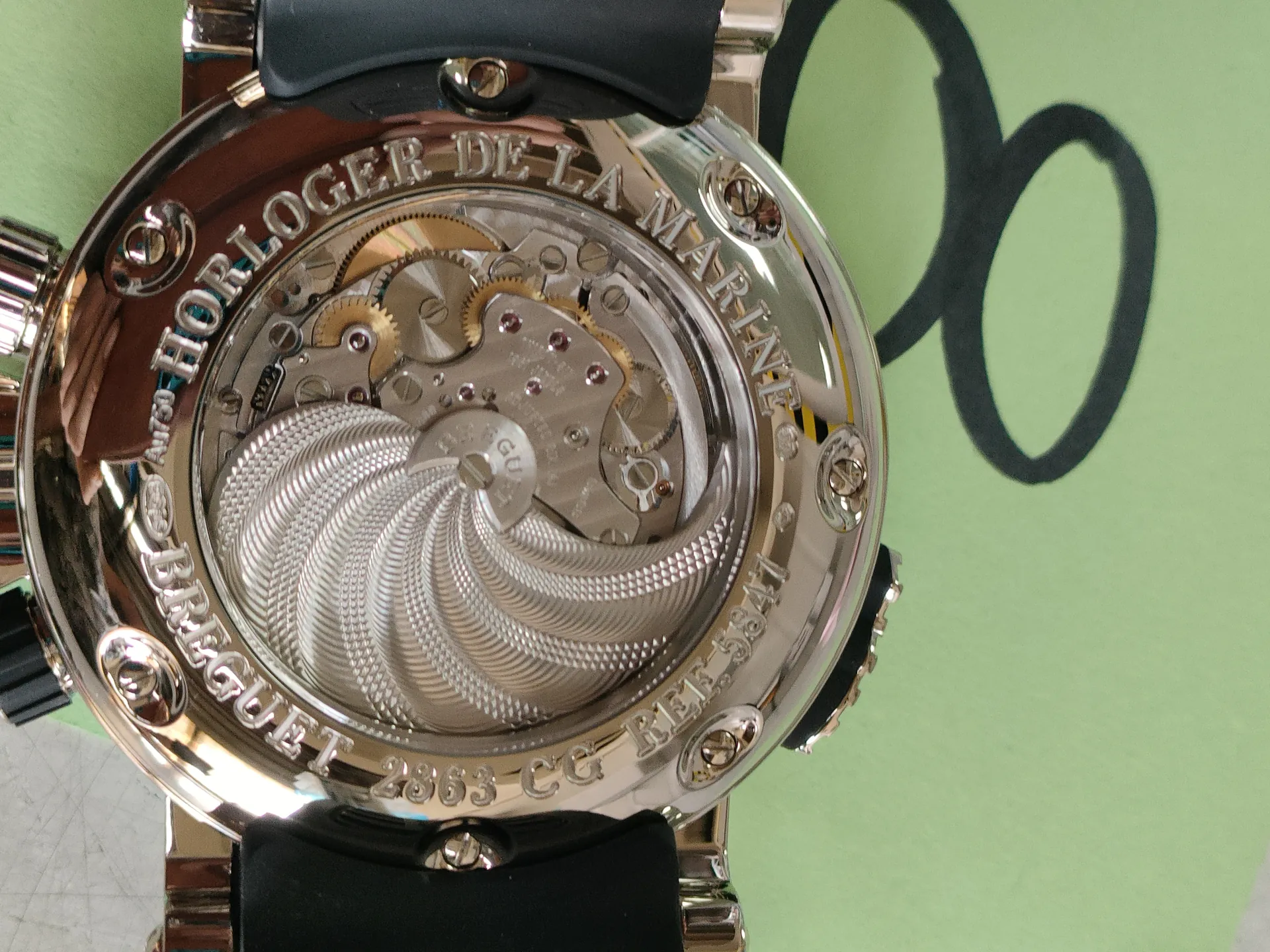 Zbliżenie na tylną część zegarka z widocznym mechanizmem. Metalowa obudowa z wygrawerowanym tekstem: HORLOGER DE LA MARINE, BREGUET 2863. Widoczne są śruby, koła zębate i spiralny wzór na tylnej pokrywie. Tło w kolorze jasnozielonym z czarnymi okrągłymi kształtami.
