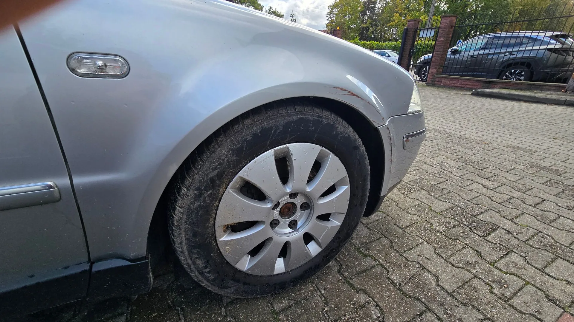 Zbliżenie na przednie lewe koło srebrnego samochodu zaparkowanego na kostce brukowej. Widoczna jest felga z plastikowym kołpakiem, opona oraz fragment przedniego błotnika i zderzaka z widocznym zarysowaniem nad nadkolem. W tle widać czarny płot i inny samochód.