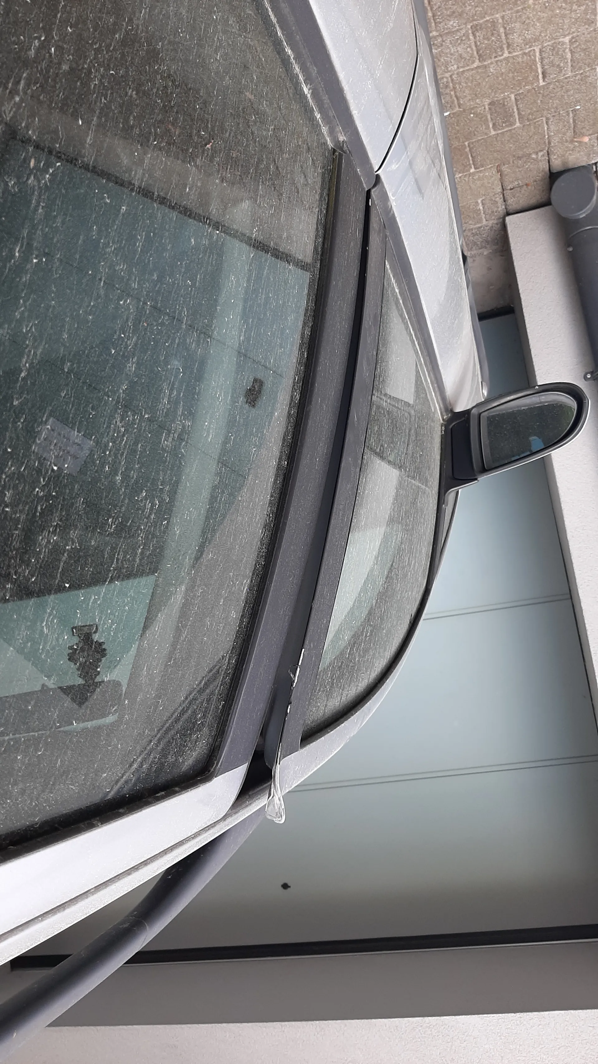 Zbliżenie na przednią szybę i fragment nadwozia srebrnego samochodu. Szyba jest zabrudzona, widoczne są ślady kurzu lub plam. Po prawej stronie obrazu znajduje się lusterko boczne. W tle biała ściana budynku i brukowana powierzchnia. Brak czytelnego tekstu.