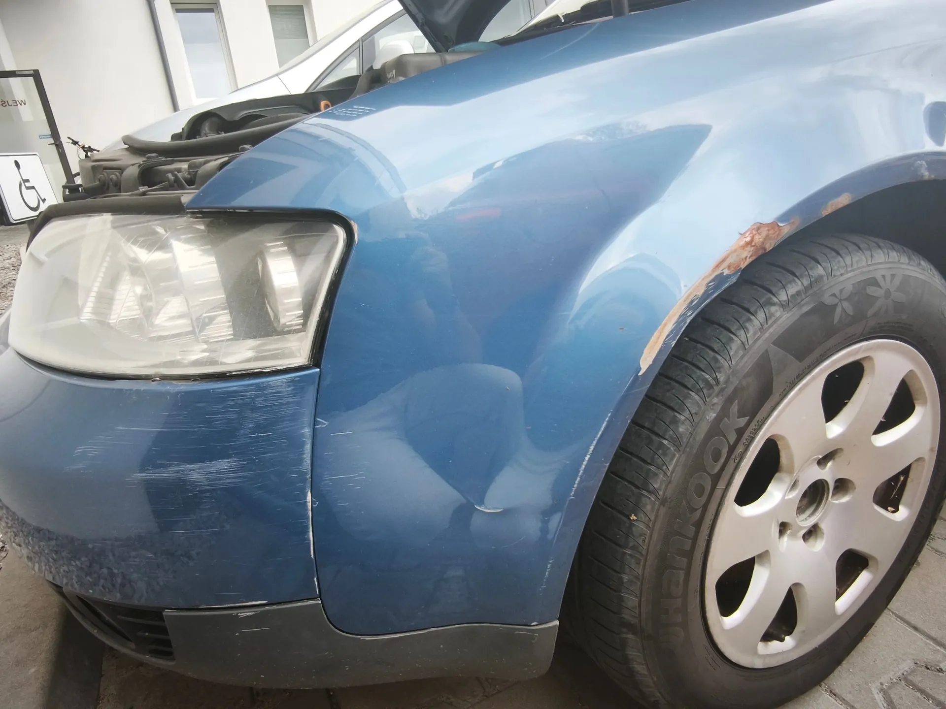 Zbliżenie na przedni lewy błotnik i reflektor niebieskiego samochodu. Na błotniku widoczne są wgniecenia oraz rdza na krawędzi nad nadkolem. Przedni zderzak posiada liczne zarysowania. Widoczna jest opona marki Hankook na srebrnej feldze aluminiowej.