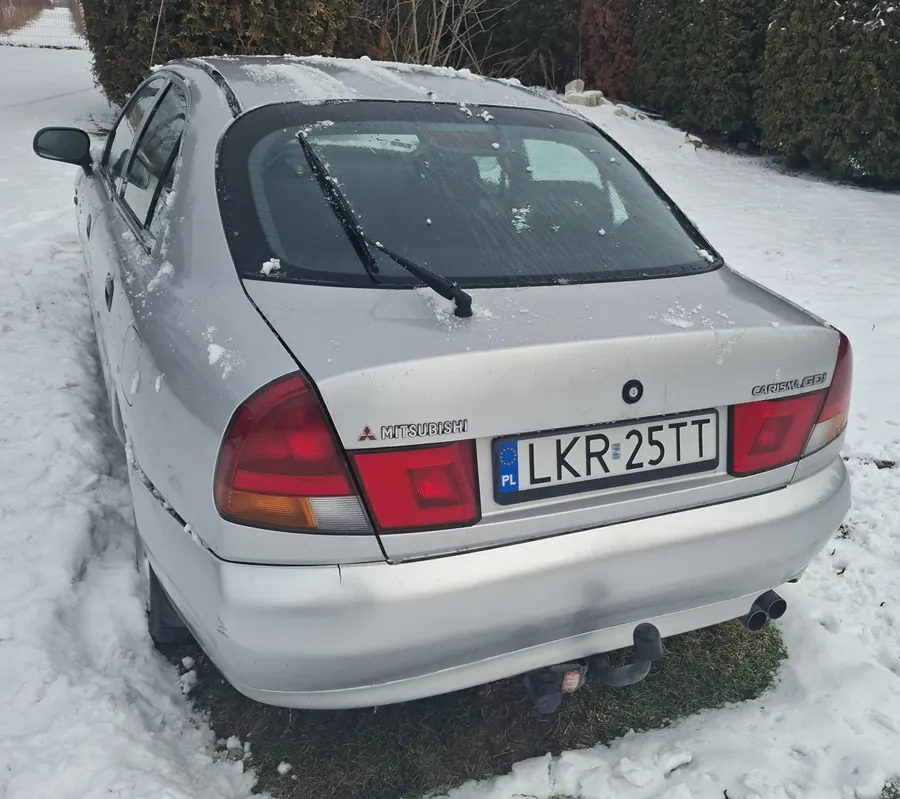 Srebrny samochód Mitsubishi z tyłu, zaparkowany na śnieżnej powierzchni. Widoczna tablica rejestracyjna PL LKR 25TT, logo Mitsubishi oraz napis CARISMA na bagażniku. Na samochodzie i otoczeniu leży śnieg, w tle krzewy i zasneżony teren. Czerwone światła tylnego oświetlenia, wycieraczka na tylnej szybie oraz dwie rury wydechowe.