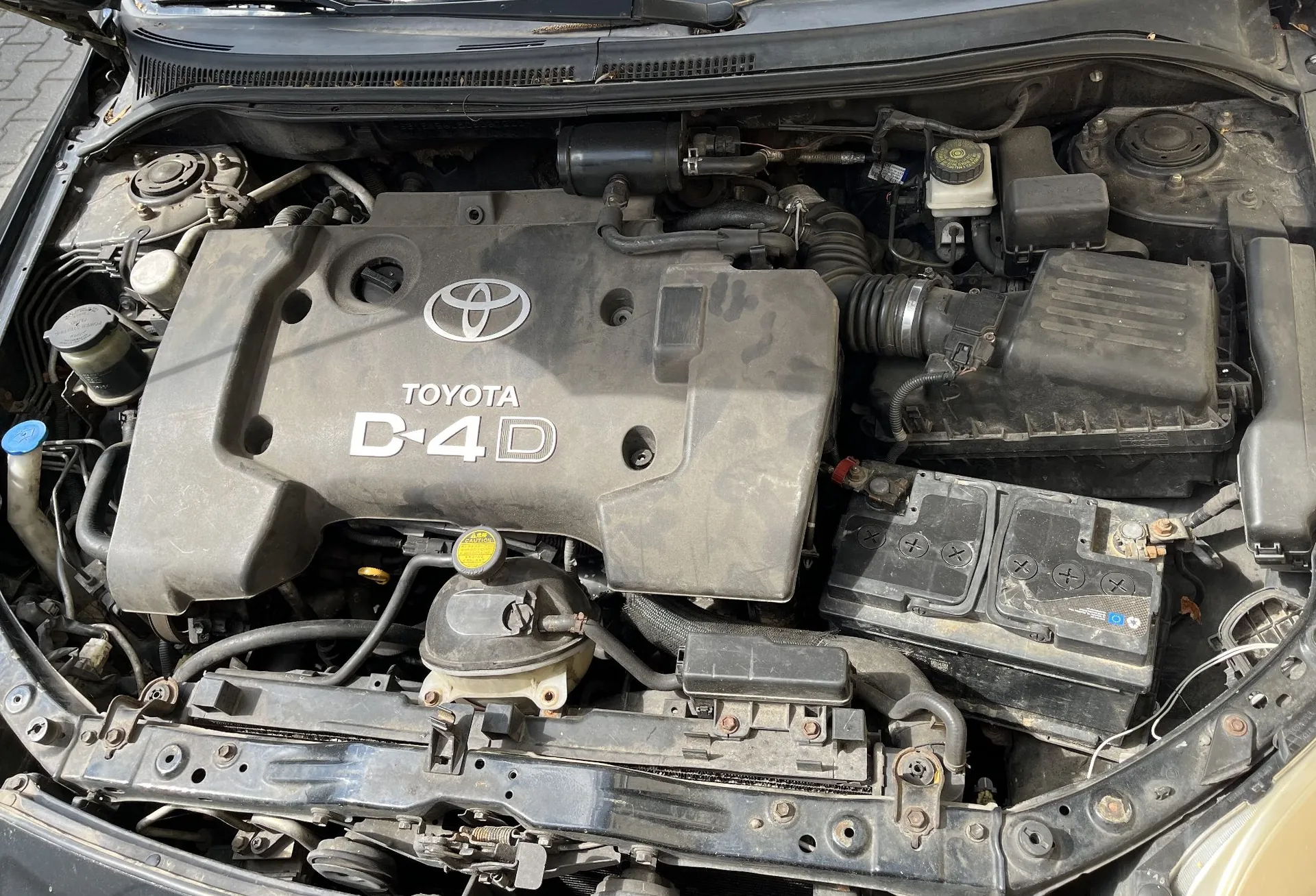 Widok z góry na komorę silnika samochodu marki Toyota. Centralnie znajduje się szara plastikowa osłona silnika z logo Toyoty oraz napisem „TOYOTA D-4D”. Po prawej stronie widoczny jest akumulator oraz obudowa filtra powietrza. Wokół silnika znajdują się różne przewody, zbiorniki z płynami i elementy mechaniczne. Całość jest zakurzona.