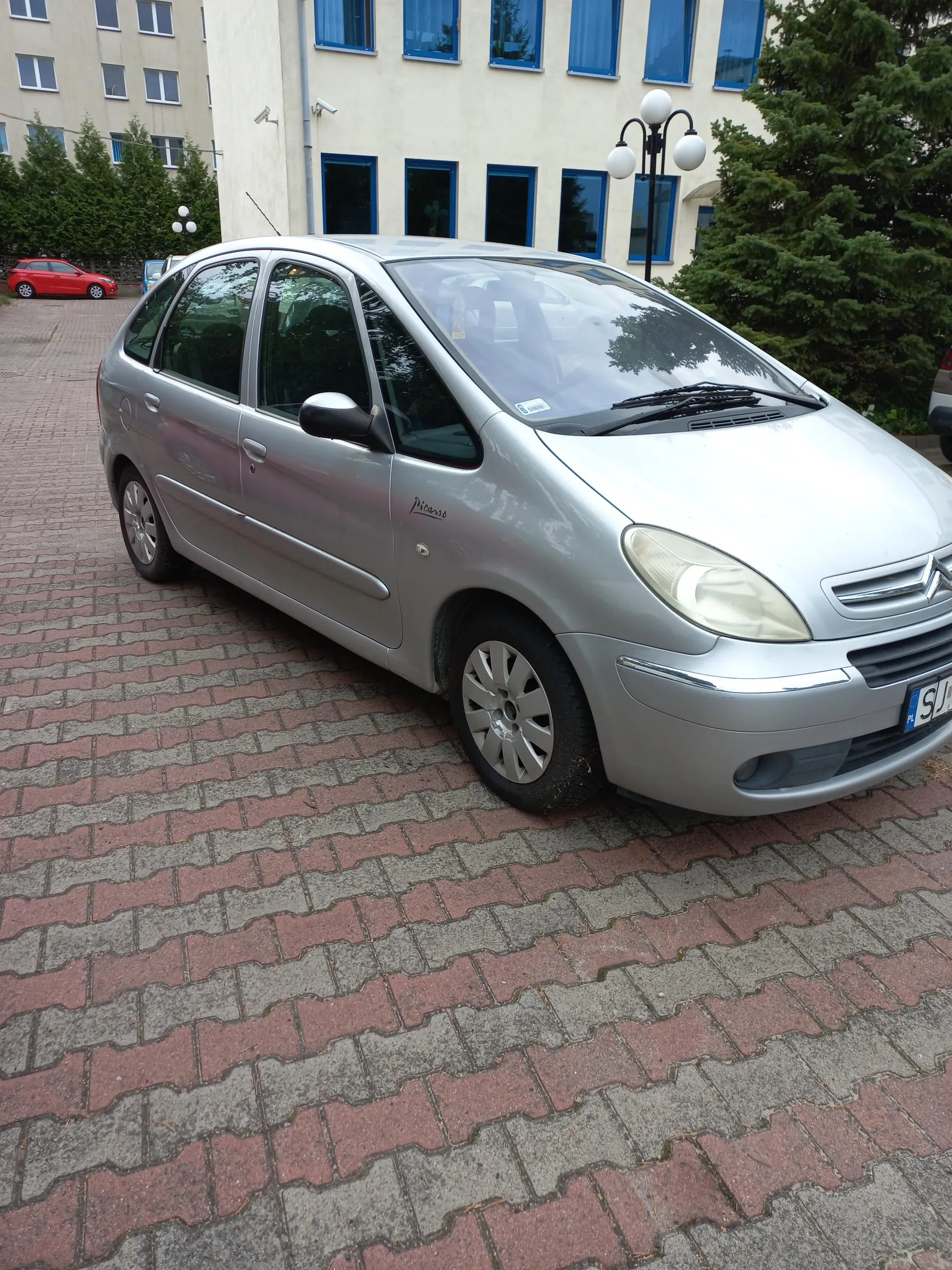Srebrny samochód osobowy marki Citroën model Xsara Picasso zaparkowany na kostce brukowej przed jasnym budynkiem z niebieskimi ramami okien. W tle widoczny jest czerwony samochód oraz zielone drzewa i krzewy.