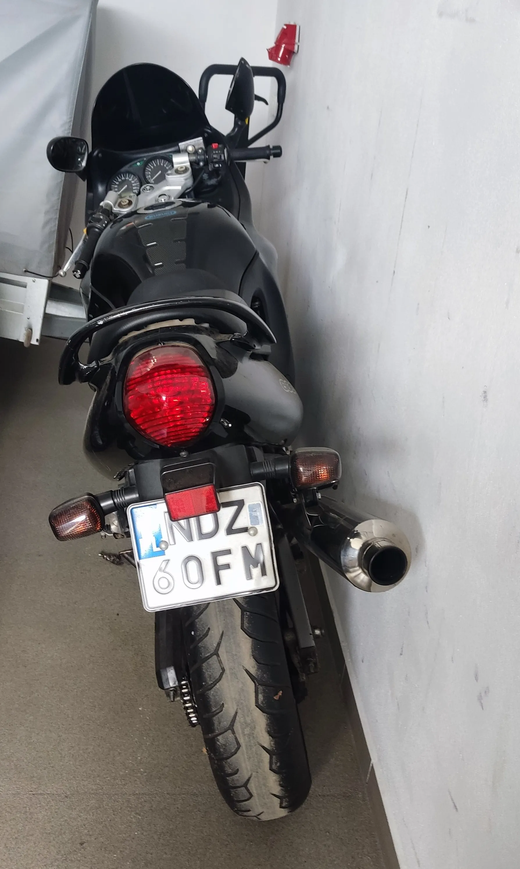 Z tyłu czarnego motocykla widoczny czerwony reflektor, tablica rejestracyjna z napisem LNDZ 60FM, chromowany wydech oraz tylne koło. Motocykl stoi przy białej ścianie, obok widoczna część szarego obiektu. Na ścianie znajduje się czerwony przedmiot.