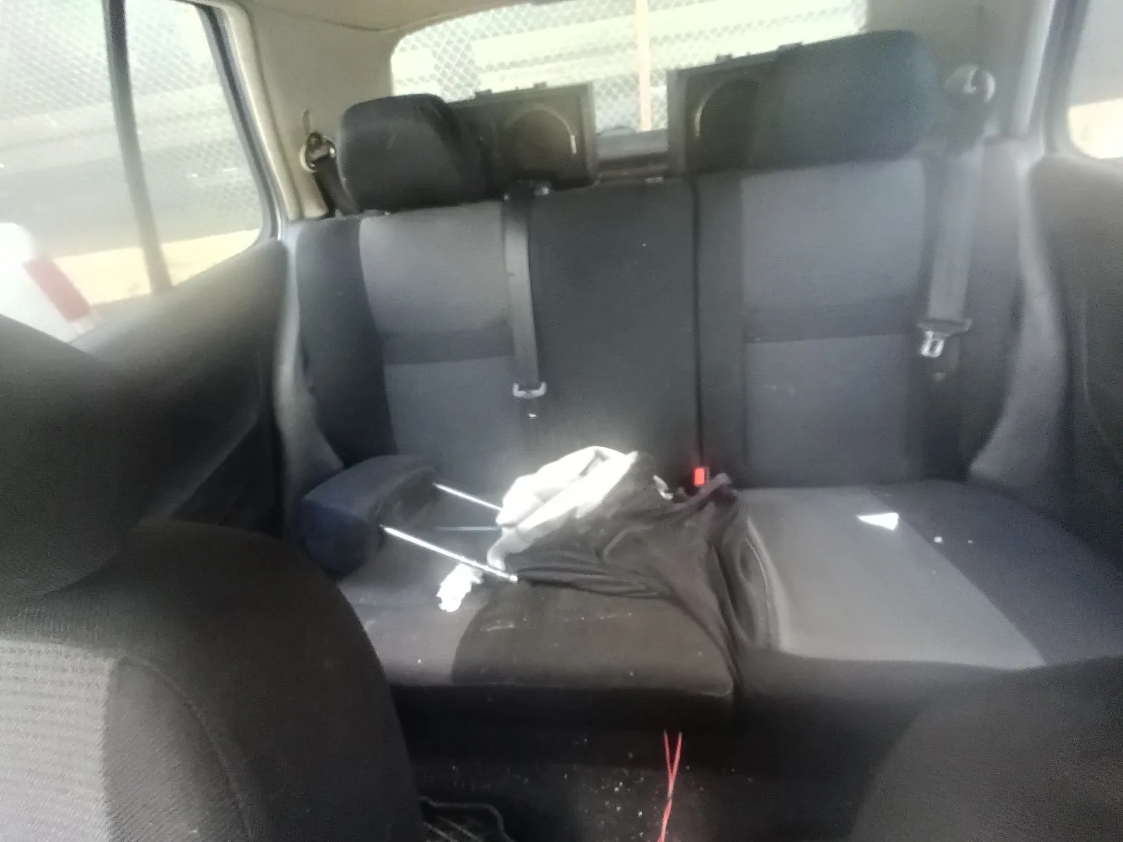 Wnętrze samochodu z widokiem na tylną kanapę w kolorze ciemnoszarym. Na siedzeniu leżą rozrzucone przedmioty: czarna tkanina oraz biały materiał. W tle widoczne są zagłówki foteli oraz tylna szyba. W lewym dolnym rogu znajduje się fragment przedniego fotela.