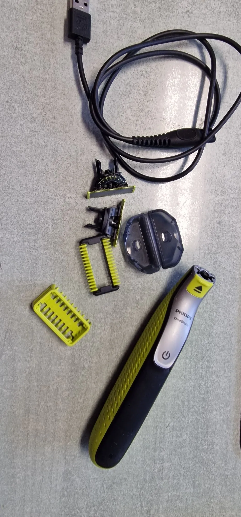 Zestaw do pielęgnacji zarostu marki Philips OneBlade rozłożony na szarym tle. W skład zestawu wchodzi czarna maszynka do golenia z limonkowymi wstawkami i napisem 'PHILIPS OneBlade', kilka wymiennych nasadek do przycinania o różnych rozmiarach (w tym jedna limonkowo-czarna i jedna w pełni limonkowa), plastikowa osłona na ostrze oraz czarny kabel USB do ładowania.