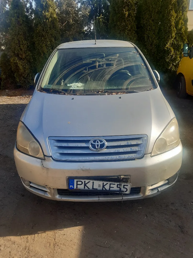 Srebrny samochód marki Toyota z przodu, z widocznym logo na atrapie chłodnicy i tablicą rejestracyjną PKL KE55. Przednia szyba i zderzak mają widoczne uszkodzenia. W tle zielone krzewy i żółty samochód.