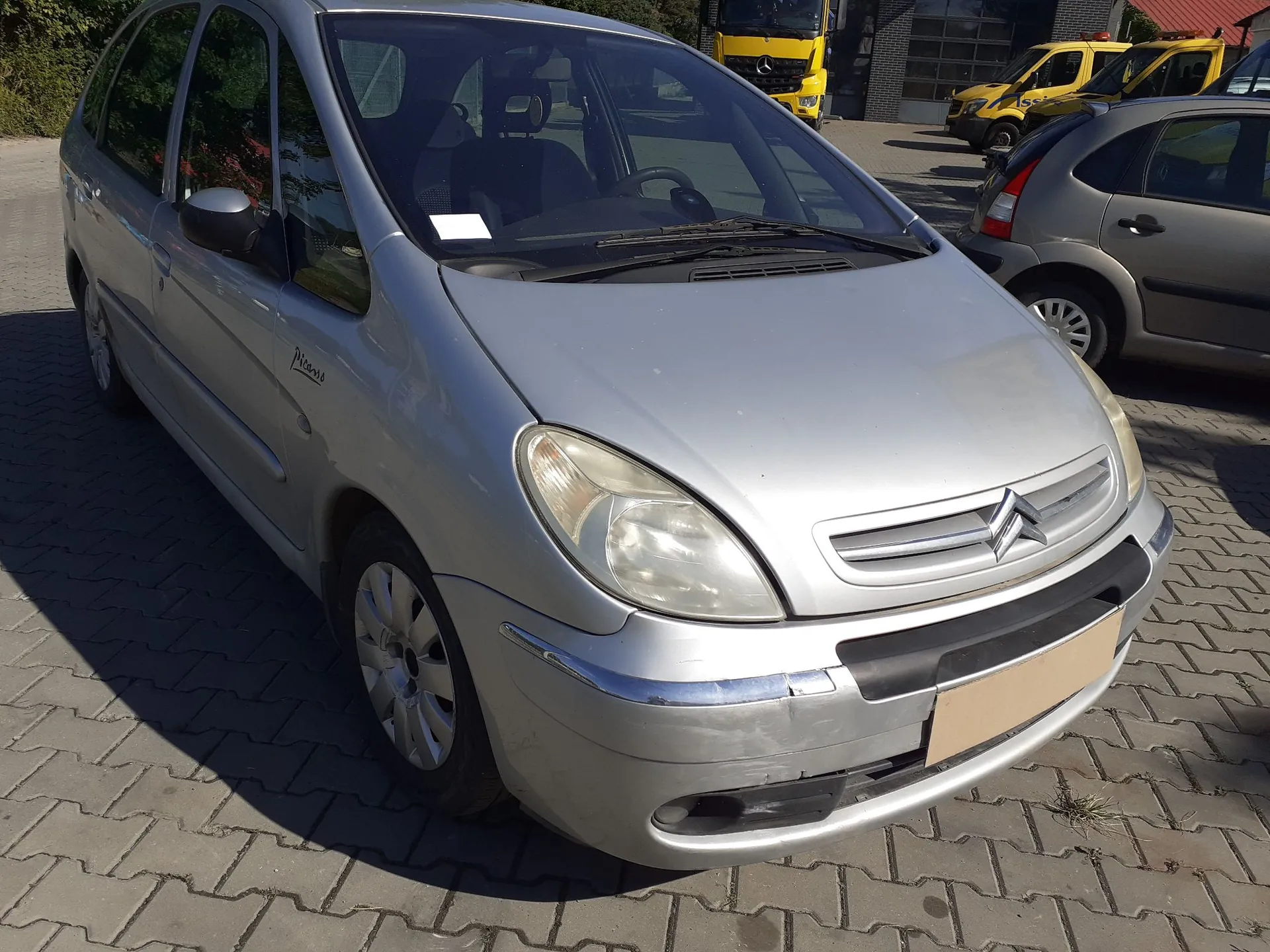 Srebrny samochód osobowy marki Citroën zaparkowany na kostce brukowej. Pojazd jest widoczny z przodu i z boku. W tle widoczne są inne samochody, w tym żółte pojazdy dostawcze oraz szary samochód osobowy.