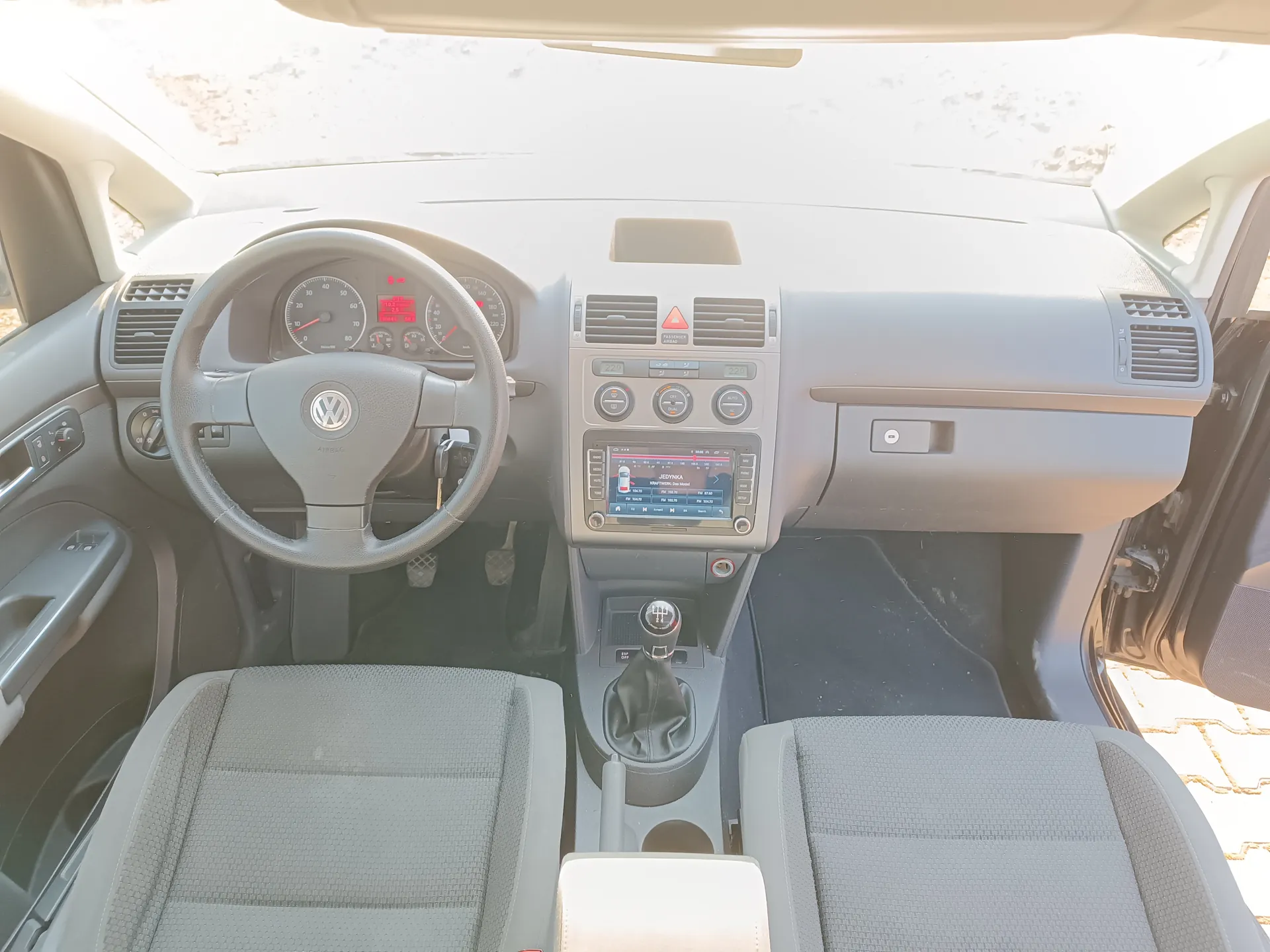 Wnętrze samochodu Volkswagen z widokiem na deskę rozdzielczą. Kierownica z logo VW, wskaźniki prędkości i obrotomierza z czerwonymi cyframi, centralny ekran multimedialny z napisem JEDYNA, klimatyzacja, dźwignia biegów, szare siedzenia tapicerowane tkaniną. Drzwi po obu stronach są otwarte, przez przednią szybę widać słoneczny dzień. Wnętrze jest jasne, z widocznymi przyciskami i wyłącznikami.