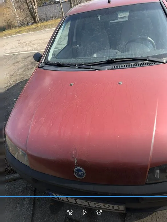 Czerwony samochód marki Fiat z widocznym logo na masce. Na masce znajdują się ślady brudu i zadrapania. Widoczna tablica rejestracyjna z częściowo przesłoniętym tekstem 'KLT S90'. Tło przedstawia parking z drzewami i drogą.