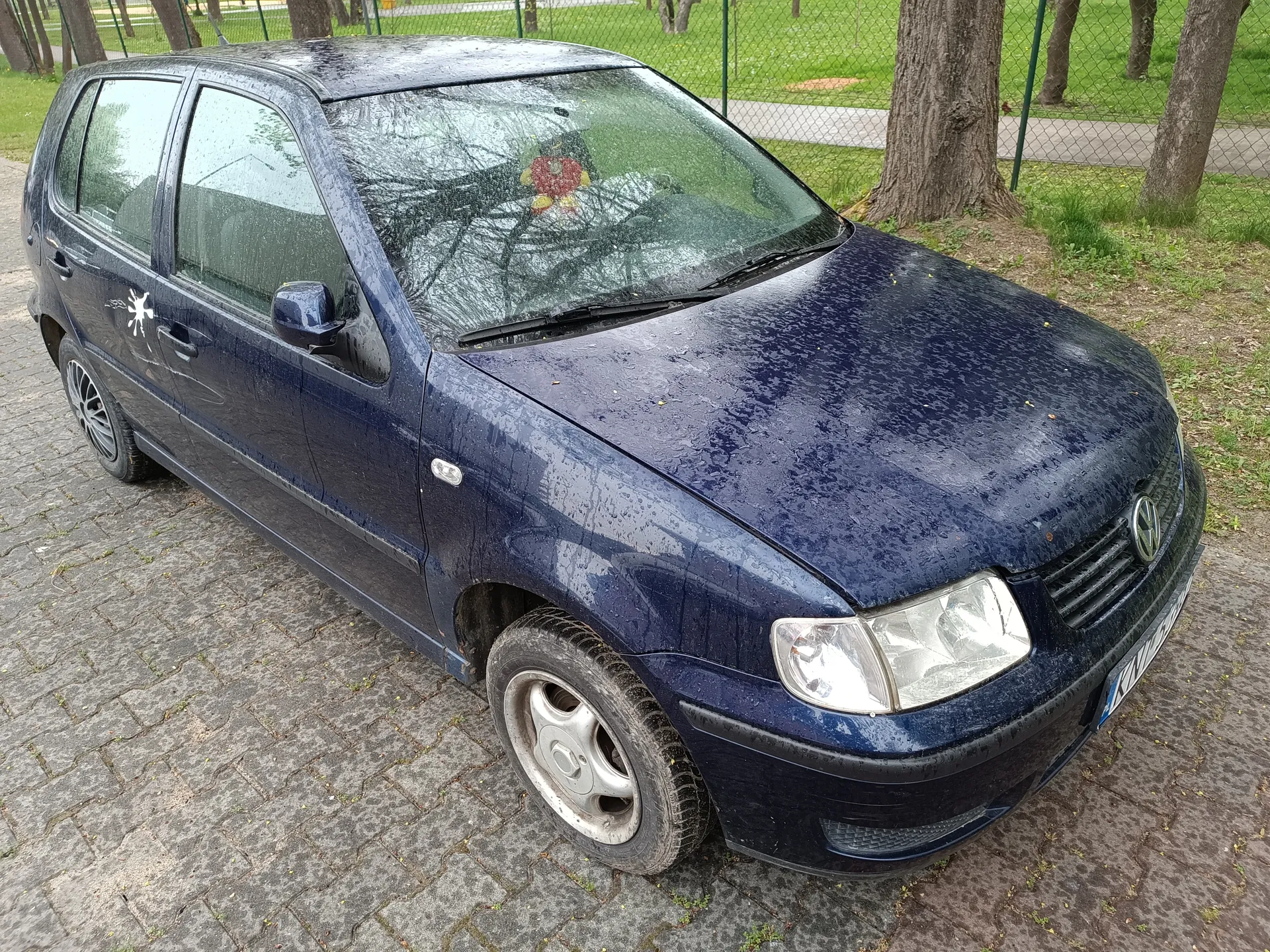 Ciemnoniebieski samochód osobowy marki Volkswagen zaparkowany na brukowanej powierzchni. Na przedniej szybie widoczny czerwono-żółty przedmiot, na drzwiach pasażerskich biały naklejony symbol przypominający gwiazdę. Samochód pokryty jest kroplami wody. W tle trawa, drzewa i siatka ogrodzeniowa. Tablica rejestracyjna na przodzie częściowo widoczna.