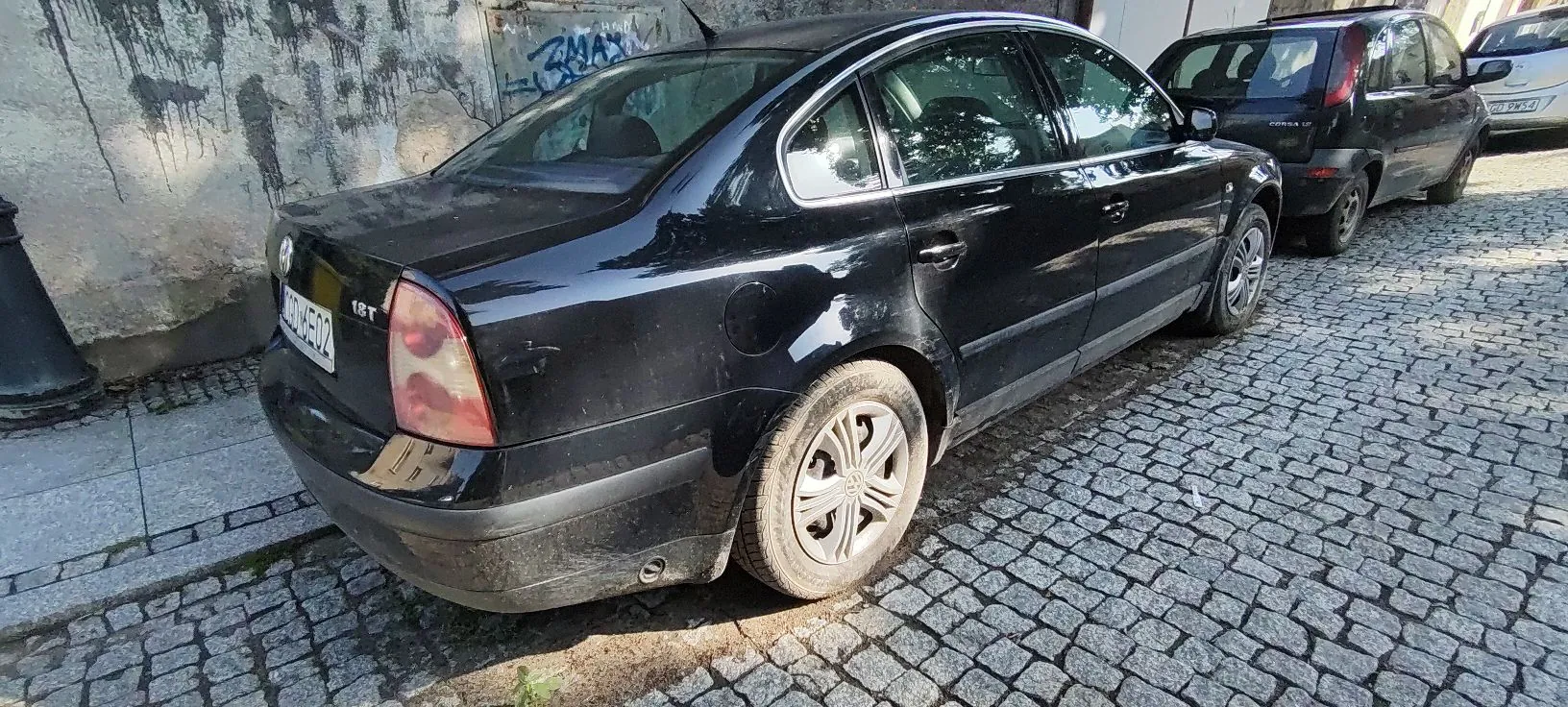 Czarny samochód osobowy zaparkowany tyłem na brukowanej ulicy obok jasnej, lekko zniszczonej ściany budynku z graffiti. Na klapie bagażnika widoczny jest napis '1.8T' oraz tablica rejestracyjna. W tle widać inny, czarny samochód zaparkowany za nim.