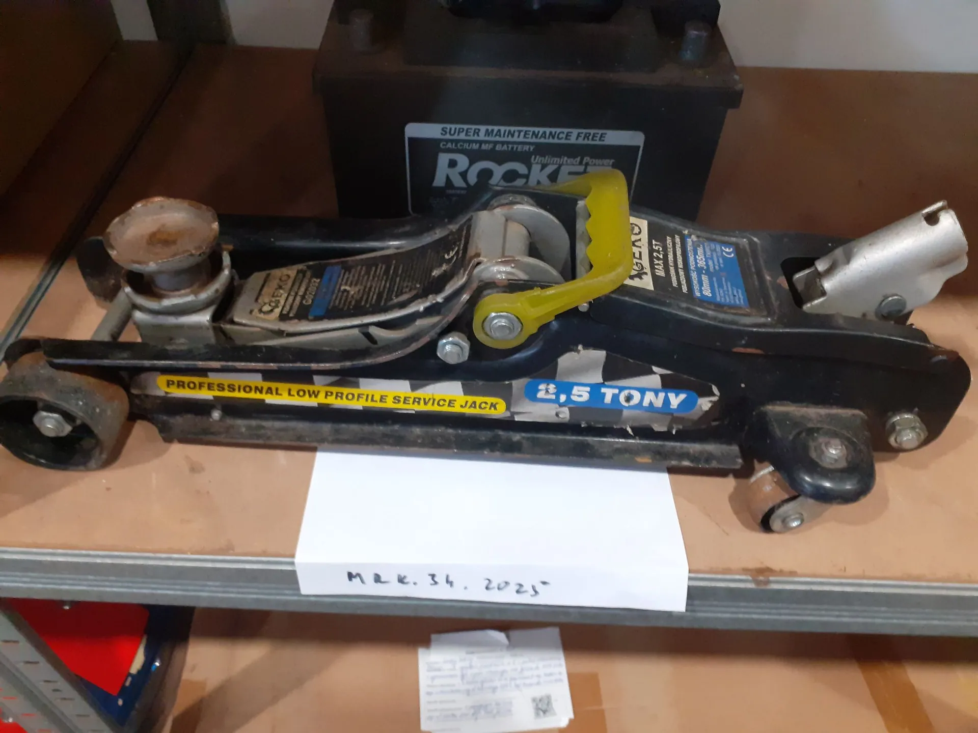Czarny podnośnik hydrauliczny o niskim profilu leżący na metalowej półce. Na boku urządzenia znajduje się żółty napis „PROFESSIONAL LOW PROFILE SERVICE JACK” oraz niebieska etykieta z napisem „2,5 TONY”. Podnośnik ma żółtą rączkę do pompowania. W tle widoczny jest czarny akumulator marki Rocket z napisem „SUPER MAINTENANCE FREE CALCIUM MF BATTERY”. Pod podnośnikiem leży biała kartka papieru z odręcznym napisem „M R K. 34. 2025”.