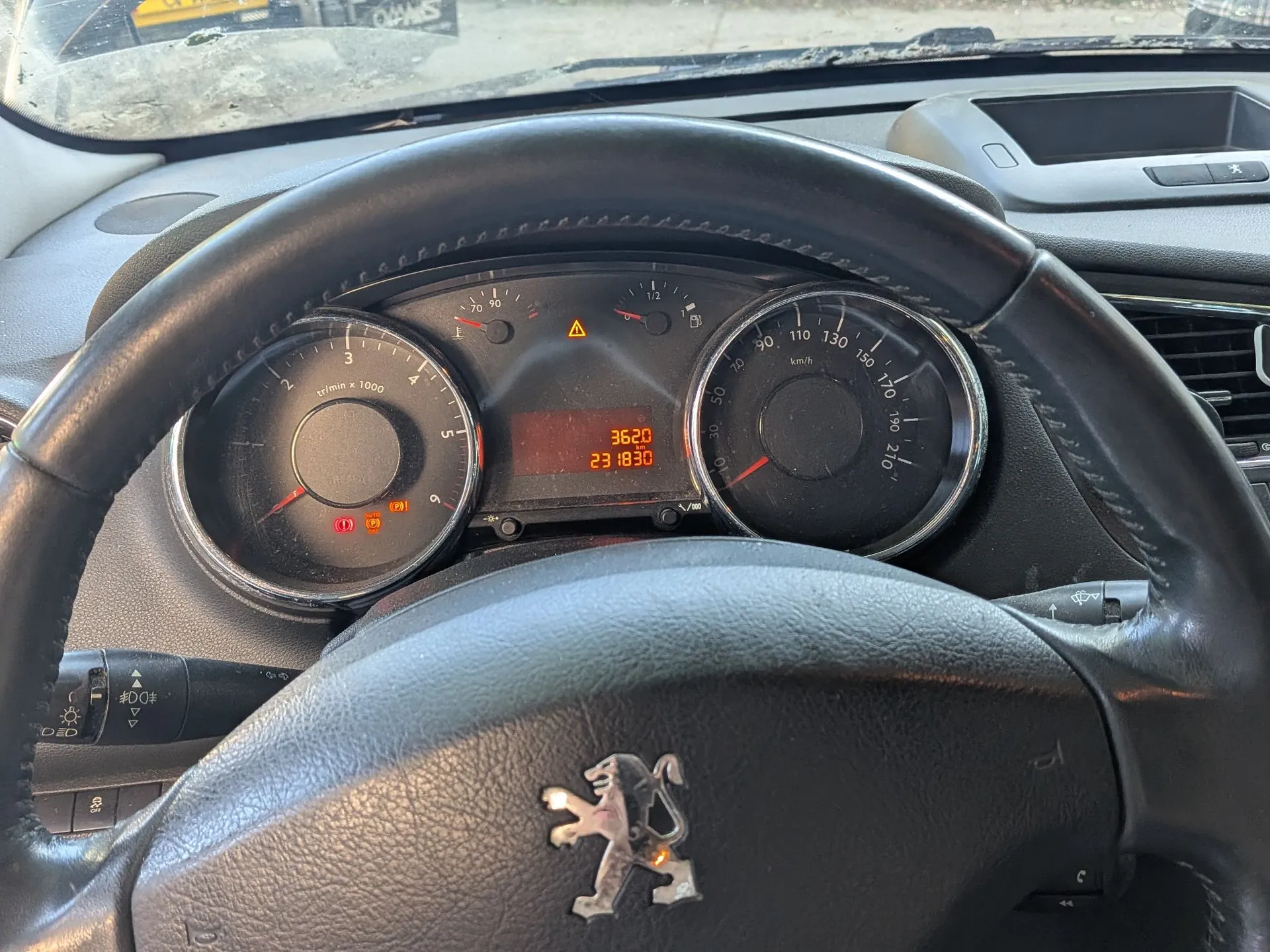 Widok z perspektywy kierowcy na wnętrze samochodu marki Peugeot. W centrum znajduje się czarna, skórzana kierownica z logo lwa na środku. Za kierownicą widoczny jest panel instrumentów z dwoma analogowymi wskaźnikami (obrotomierzem po lewej i prędkościomierzem po prawej) oraz czerwonym wyświetlaczem cyfrowym w środku, na którym widnieją liczby 3620 oraz 23 1830. Na desce rozdzielczej widoczne są kontrolki ostrzegawcze.