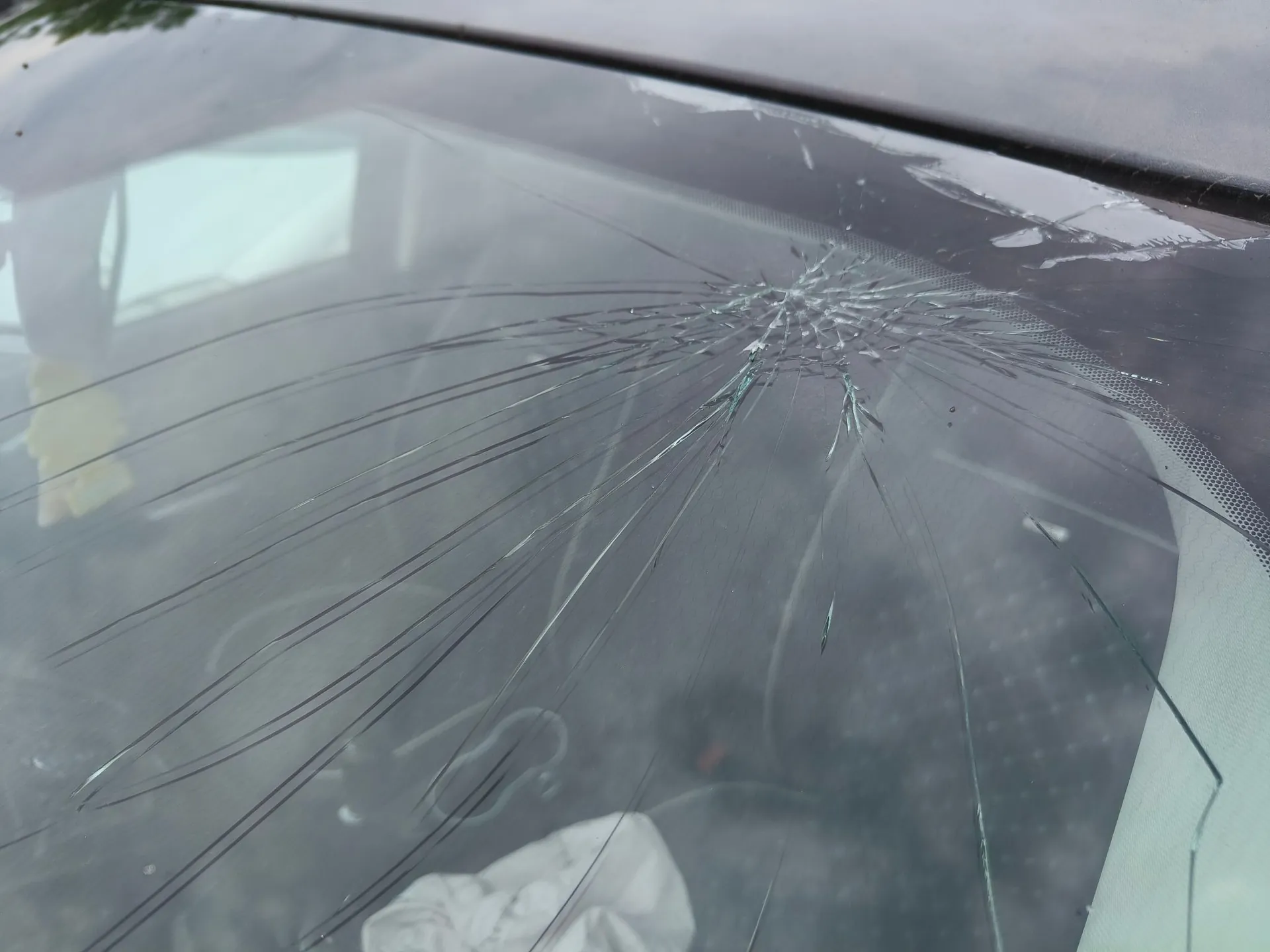 Zbliżenie na pękniętą przednią szybę samochodu. W górnej części szyby widoczne jest rozległe rozbicie w kształcie pajęczyny z licznymi pęknięciami rozchodzącymi się promieniście w dół i na boki. W tle, za szybą, widoczne jest wnętrze pojazdu.