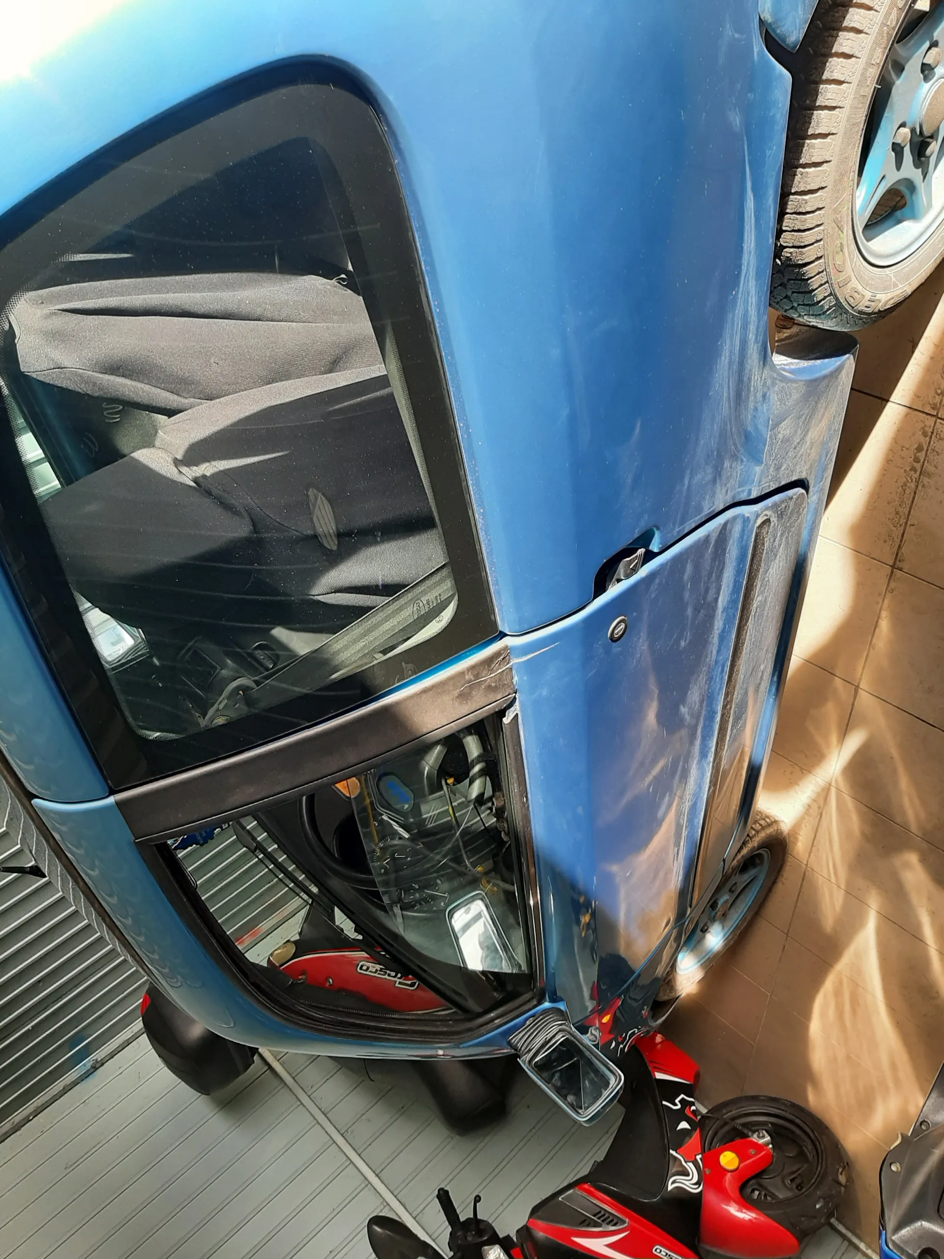 Niebieski samochód z otwartą drzwią, widoczne wnętrze z szarymi siedzeniami. Obok niebieskiego samochodu czerwony skuter z białymi wzorami. Podłoga z kafelkami, w tle metalowa brama garażowa.