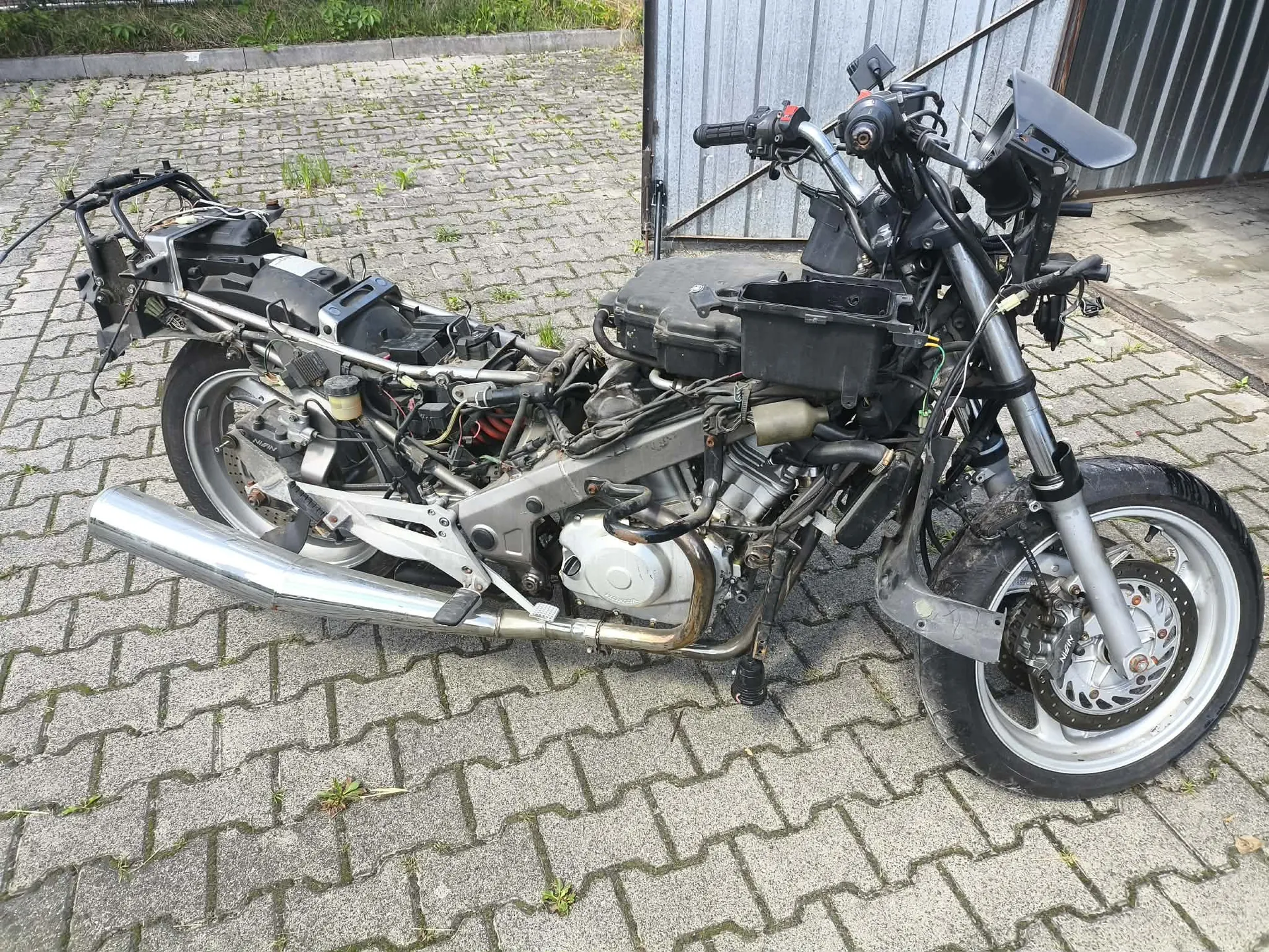 Zdjęcie czarnego motocykla w stanie rozbiórki, pozbawionego owiewek i wielu elementów nadwozia, stojącego na kostce brukowej przed szarym budynkiem. Widoczna jest rama, silnik, srebrna rura wydechowa oraz przednie i tylne koła.