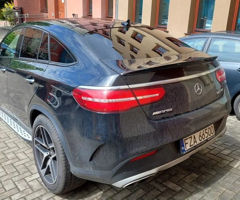 Czarny samochód Mercedes-AMG z tyłu, widoczny numer rejestracyjny FZA 66500 z oznaczeniem PL, czerwone światła tylnego, emblemat AMG i logo Mercedesa. Samochód zaparkowany na brukowanej powierzchni, w tle budynki z oknami i inny pojazd.