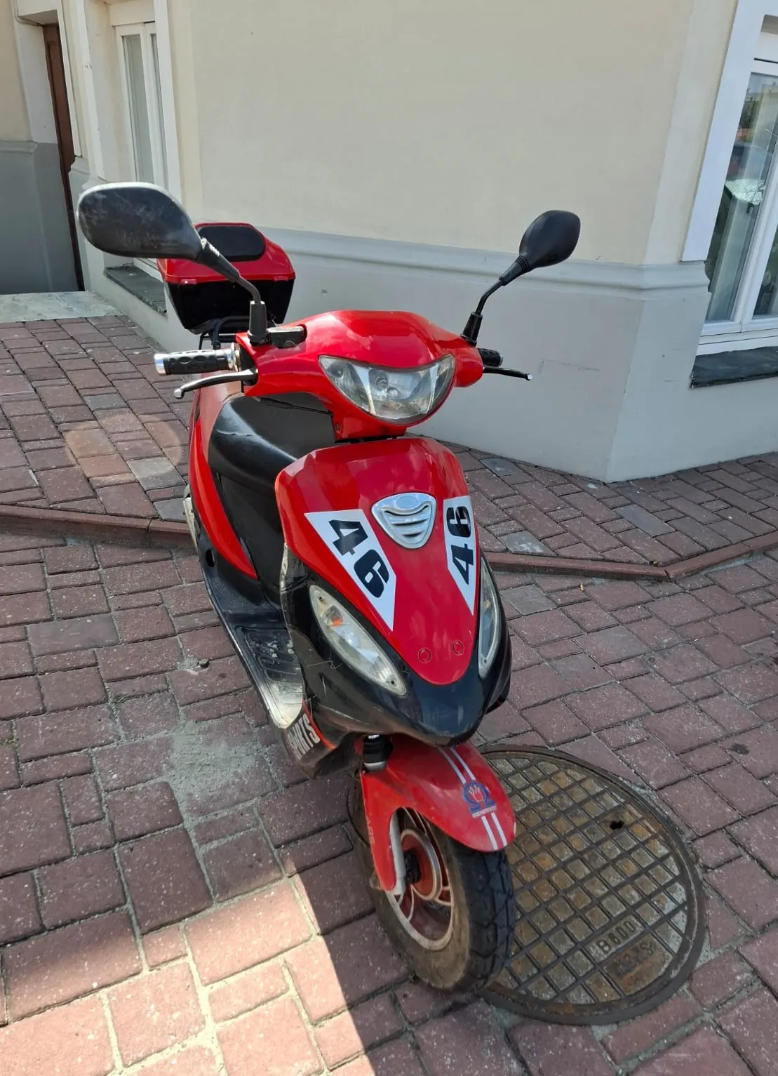 Czerwony skuter zaparkowany na chodniku z czerwonej kostki brukowej, obok metalowego włazu kanalizacyjnego. Skuter posiada czarne elementy plastikowe oraz białe naklejki z numerem '46' na przedniej owiewce. W tle widoczna jest jasna ściana budynku z oknami.