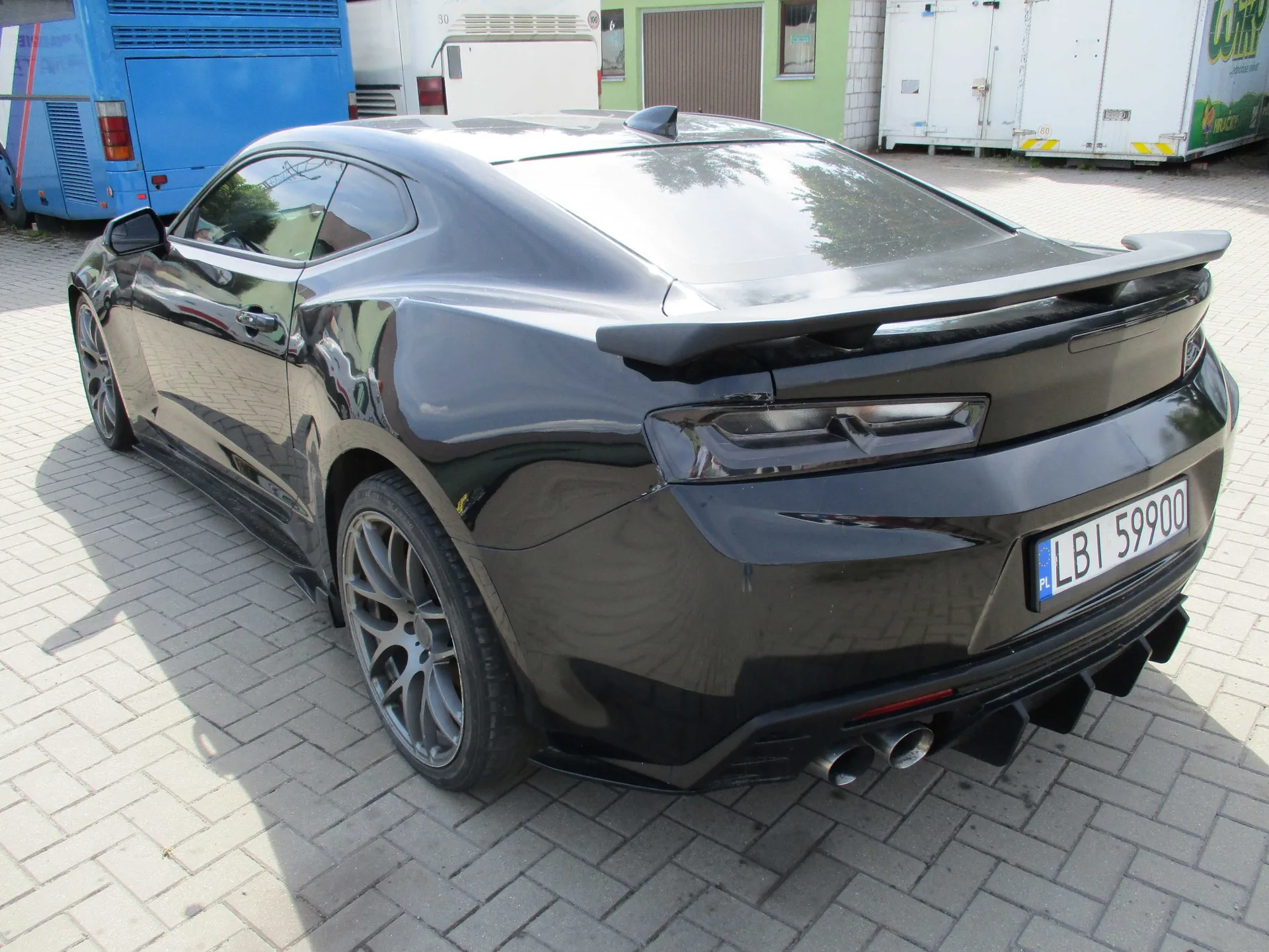 Czarny samochód sportowy typu coupe zaparkowany na kostce brukowej. Widok z tyłu i z boku. Pojazd posiada spojler na klapie bagażnika, ciemne felgi oraz tablicę rejestracyjną o treści LBI 59900. W tle widoczne są białe i niebieskie furgonetki oraz fragment budynku z brązową bramą garażową.