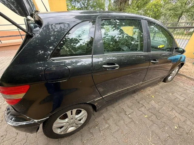 Boczny widok czarnego samochodu typu hatchback zaparkowanego na kostce brukowej. Widoczna jest prawa strona pojazdu z dwiema drzwiami, otwartą klapą bagażnika oraz srebrnymi felgami aluminiowymi. W tle widać żółtą ścianę budynku oraz zielone drzewa.