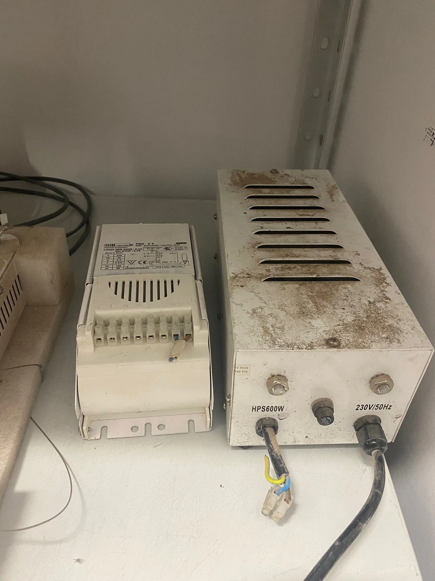 Dwa białe urządzenia elektroniczne umieszczone na białej powierzchni. Po lewej stronie mniejszy moduł z widocznymi złączami i częściowo czytelną etykietą z symbolami. Po prawej większy, zabrudzony brązowymi plamami aparat z poziomymi szczelinami wentylacyjnymi na górze. Na jego przedniej części widoczne napisy: 'HPS600W' oraz '230V/50Hz'. Do urządzenia podłączone są przewody, w tym czarny kabel i odsłonięte przewody żółty, niebieski i biały. W tle biała ściana i czarne przewody po lewej stronie.
