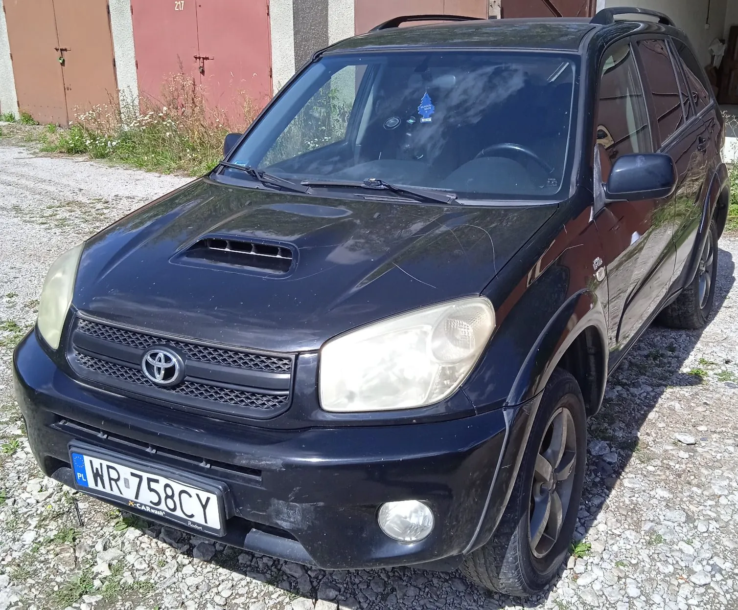 Czarny samochód marki Toyota z nadbudową typu SUV, zaparkowany na żwirowym podłożu przed czerwonymi drzwiami garażowymi. Pojazd posiada maskę z wlotem powietrza, polską tablicę rejestracyjną o numerze WR 758CY oraz srebrne logo Toyoty na grillu.