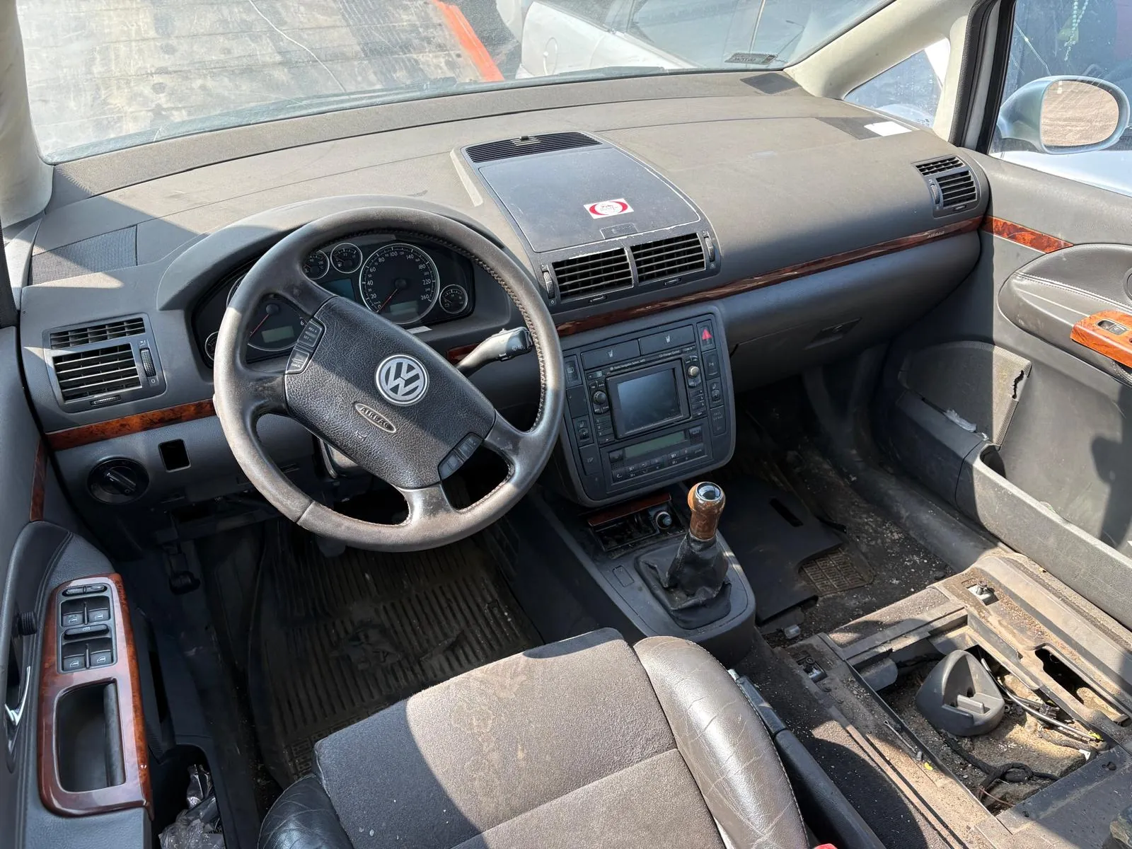 Wnętrze samochodu Volkswagen z widoczną kierownicą z logo VW, tablicą przyrządów z licznikami, centralnym panelem z ekranem i przyciskami, dźwignią zmiany biegów z drewnianą rączką oraz brudnymi siedzeniami. Deska rozdzielcza posiada brązowe wstęgi dekoracyjne, a na niej naklejkę z czerwonym symbolem. W tle przez przednią szybę widoczne są inne pojazdy. Wnętrze wydaje się zużyte, z widocznymi zabrudzeniami i częściowo rozebranym elementem po prawej stronie.
