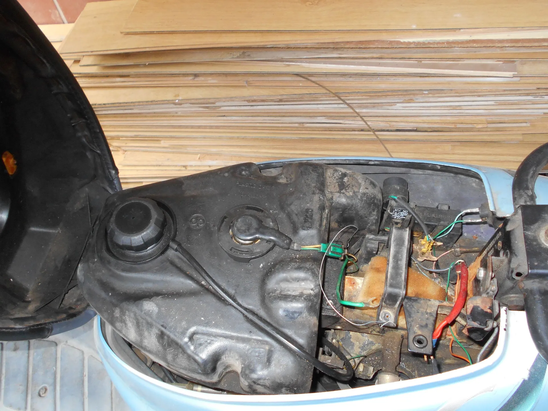 Widok z góry na wnętrze nadwozia jasnoniebieskiego skutera. Widoczny jest czarny, plastikowy zbiornik paliwa z zakrętką oraz system przewodów elektrycznych i rurek. W tle znajduje się stos jasnych, drewnianych płyt. Po lewej stronie widoczny jest fragment czarnego elementu plastikowego.