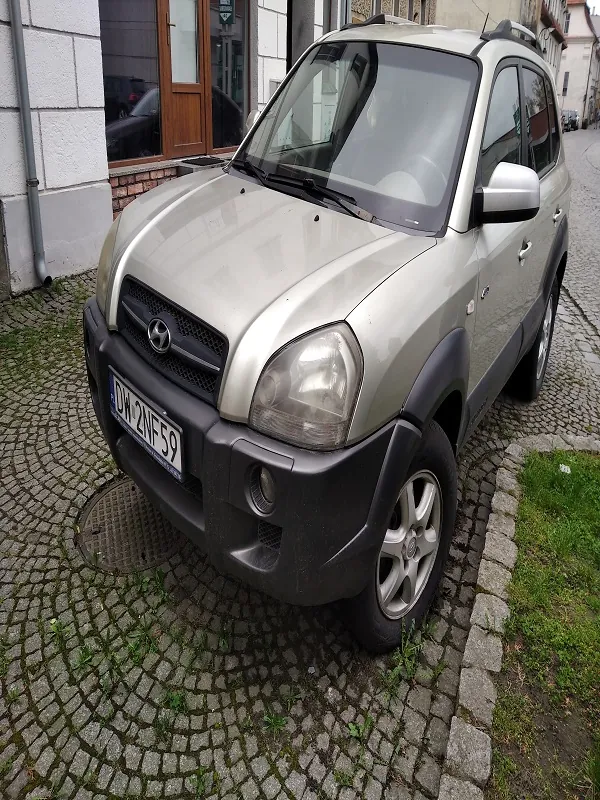 Srebrny samochód typu SUV marki Hyundai zaparkowany na brukowanej ulicy. Widoczna tablica rejestracyjna z numerem DW 2MF59. Po lewej stronie budynek z białymi płytkami i brązowymi drzwiami, po prawej trawa między kamieniami. Tło z budynkami i częściowo widocznym samochodem w oddali.