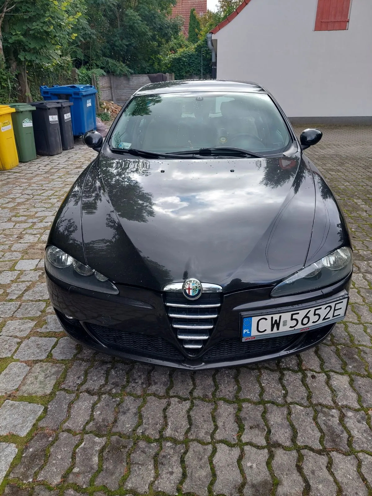Czarny samochód marki Alfa Romeo zaparkowany przodem do obiektywu na kostce brukowej. Na masce samochodu odbijają się chmury i drzewa. Pojazd posiada polską tablicę rejestracyjną o numerze CW 5652E. W tle widoczne są kolorowe kontenery na odpady (żółty, zielony, szary, niebieski), zieleń oraz fragment białego budynku z czerwonymi okiennicami.