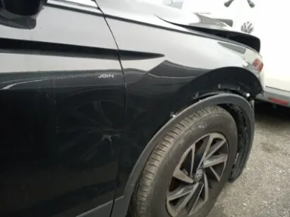 Czarny samochód widziany z boku od tyłu, z widocznym kołem z aluminiową felgą, fragmentem tylnego światła i napisem "JOIN" na boku nad kołem. W tle częściowo widoczny biały pojazd oraz nawierzchnia z żwiru.