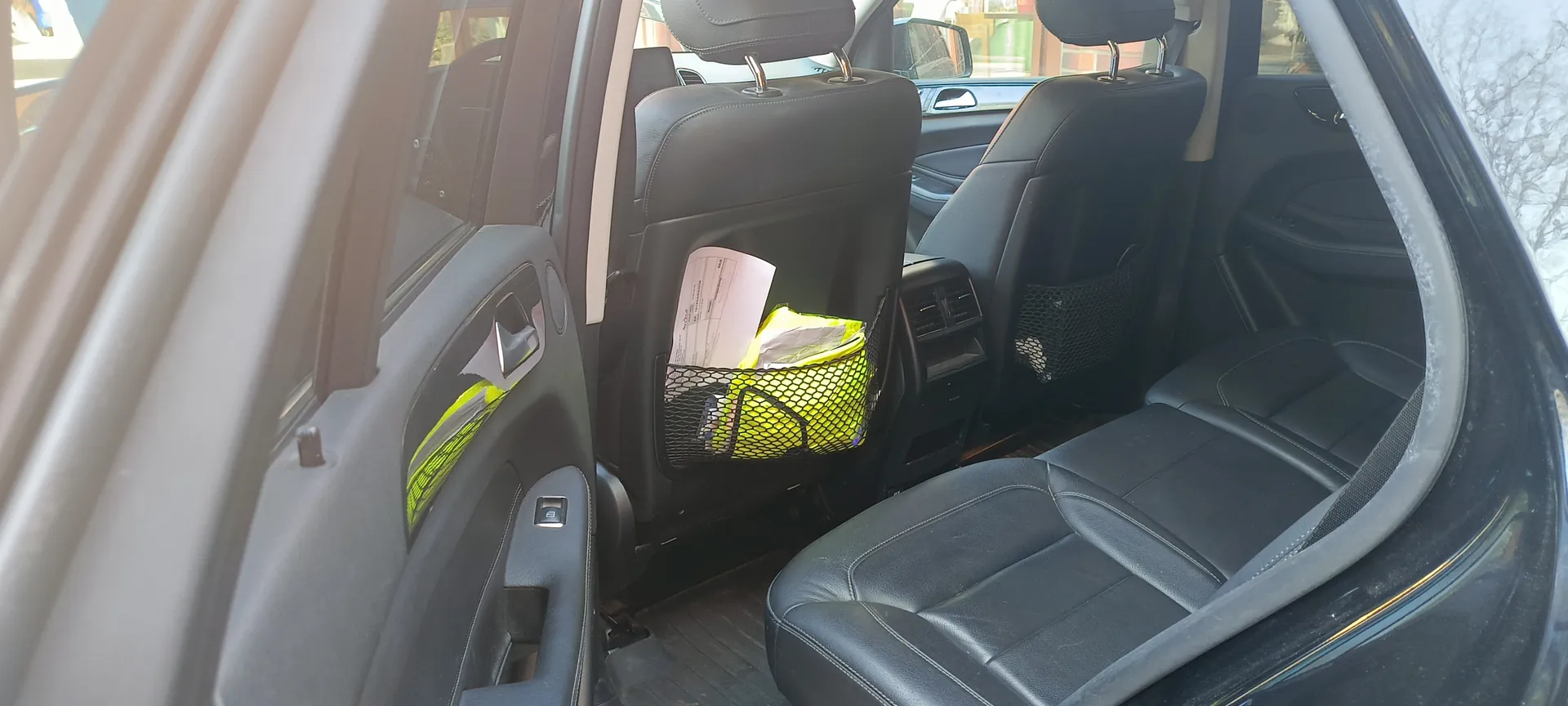 Wnętrze samochodu z widocznymi czarnymi skórzonymi fotelami. Drzwi po lewej stronie są otwarte, a na oparciach przednich foteli umieszczono siateczki. W siateczce po lewej stronie znajduje się żółta torba i białe dokumenty. W tle przez przednią szybę widać fragment budynku i drzewa. Światło słoneczne wpada przez okno, tworząc połysk na lewej stronie obrazu.