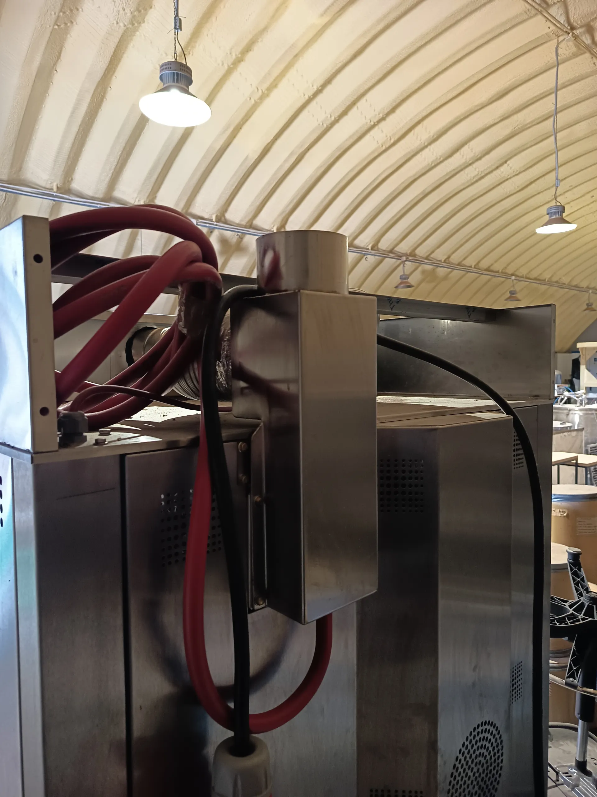 Przemysłowe urządzenia ze stali nierdzewnej z czerwonymi i czarnymi wężami, pod sufitem o łukowym kształcie z żebrowaną strukturą i wiszącymi lampami. W tle widoczne są dodatkowe urządzenia i beczki.