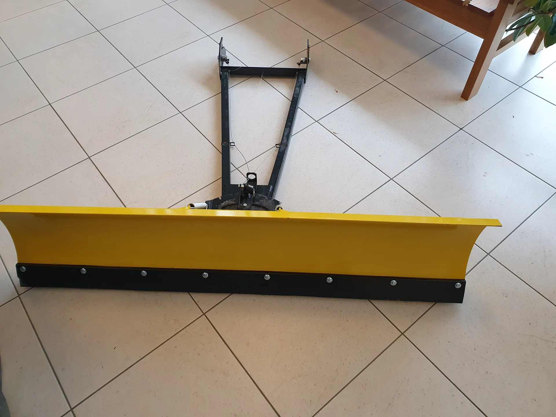 Żółty pług śnieżny z czarną podstawą i metalowym ramieniem umieszczony na podłodze z jasnymi płytkami. W tle widoczne są nogi drewnianego mebla i zielone liście rośliny.