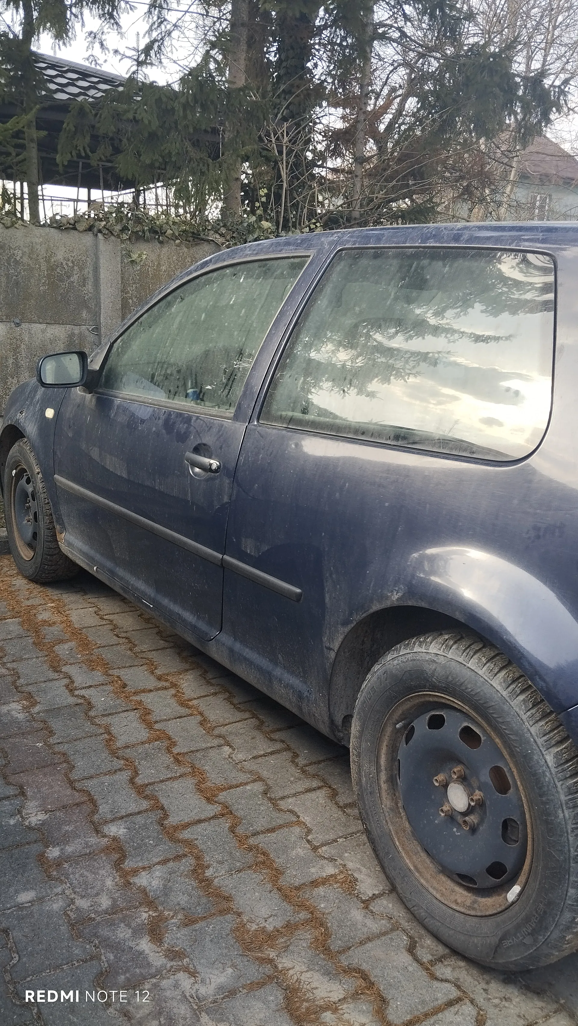 Niebieski samochód osobowy zaparkowany na brukowanej powierzchni. Samochód jest zabrudzony, widoczne są brudne opony i karoseria. W tle betonowa ściana z pnączami, drzewa iglaste i liściaste oraz fragment budynku z dachem. Na dole zdjęcia widoczny napis 'REDMI NOTE 12'.